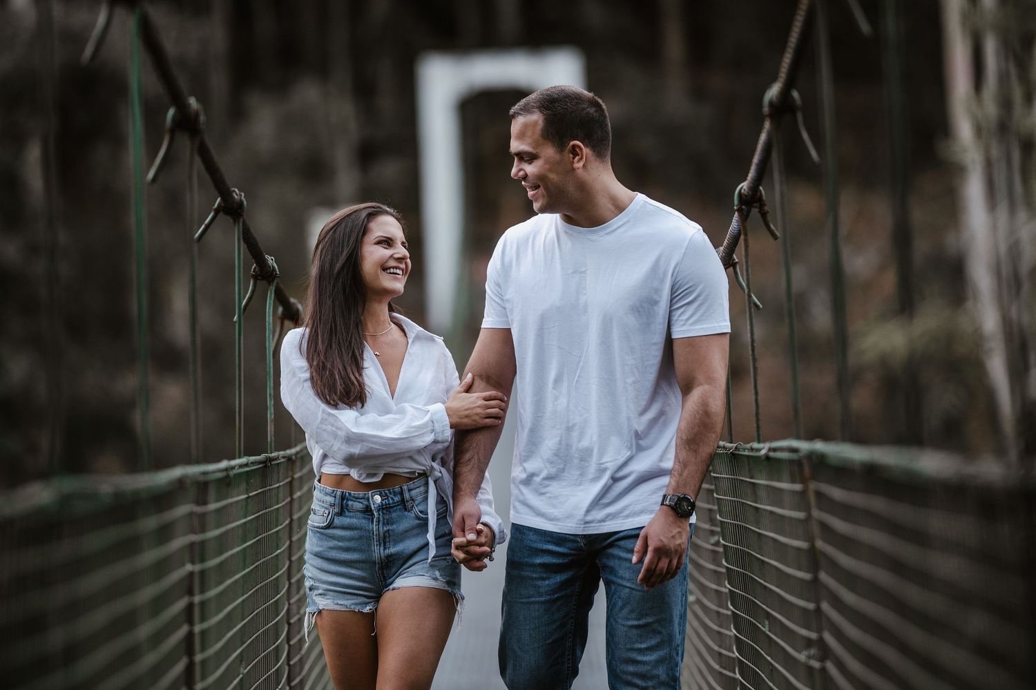 Couple walking on a wooden suspension bridge in casual summer outfits while holding hands.