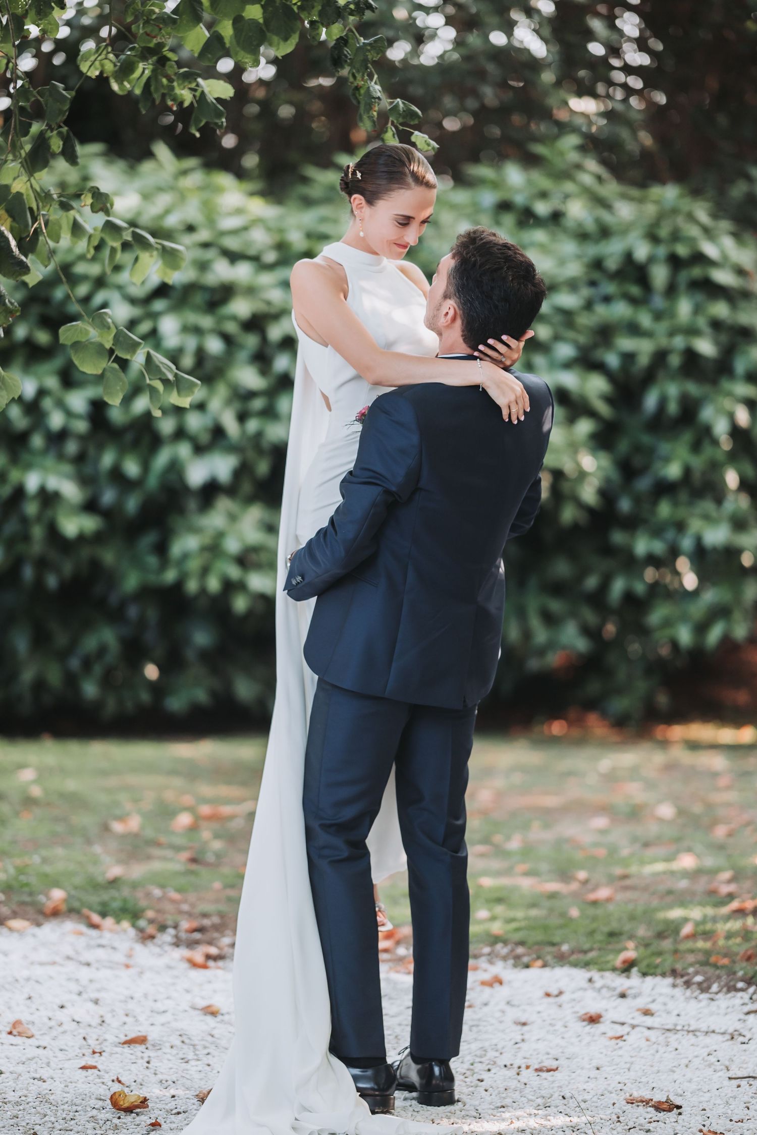 Newlyweds embrace on a gravel path surrounded by lush green foliage during their outdoor wedding photoshoot.