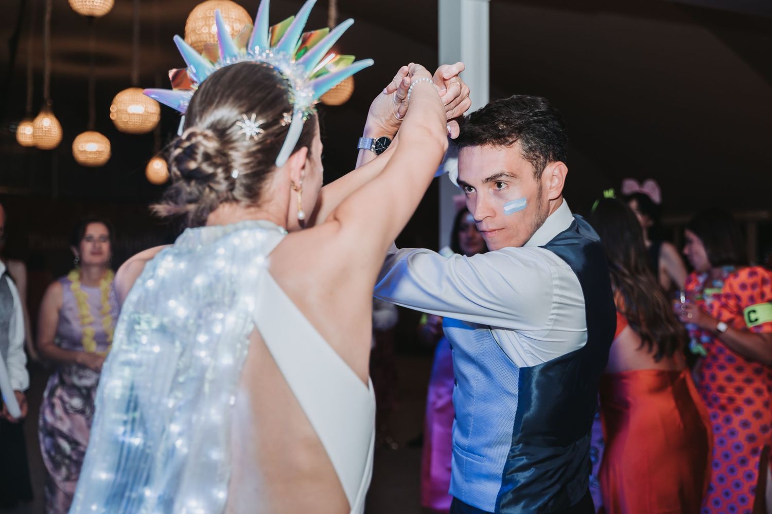 A couple dances together on the dance floor at a wedding reception with soft lighting and other guests in the background.