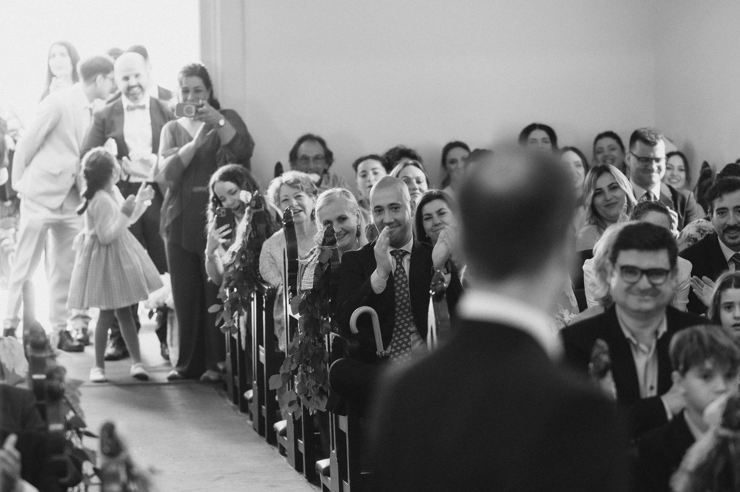 Wedding ceremony guests seated in rows with natural light streaming through windows.