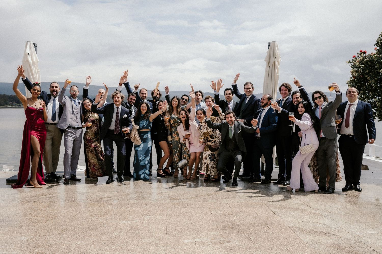 Large group of wedding guests arranged in rows on a beach for a panoramic photo with arms raised in celebration.
