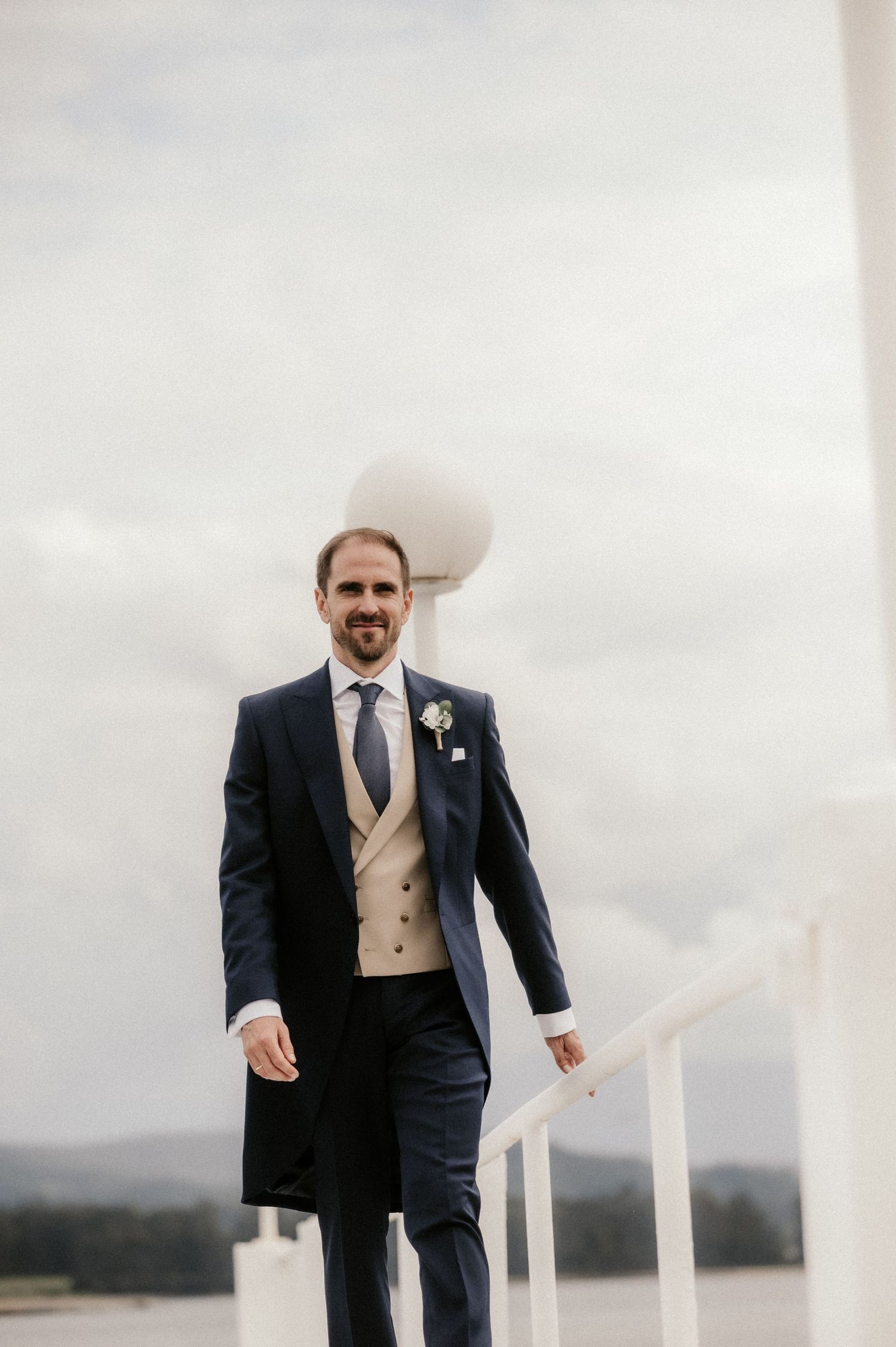A groom in a navy suit and tan vest stands against a cloudy sky backdrop.