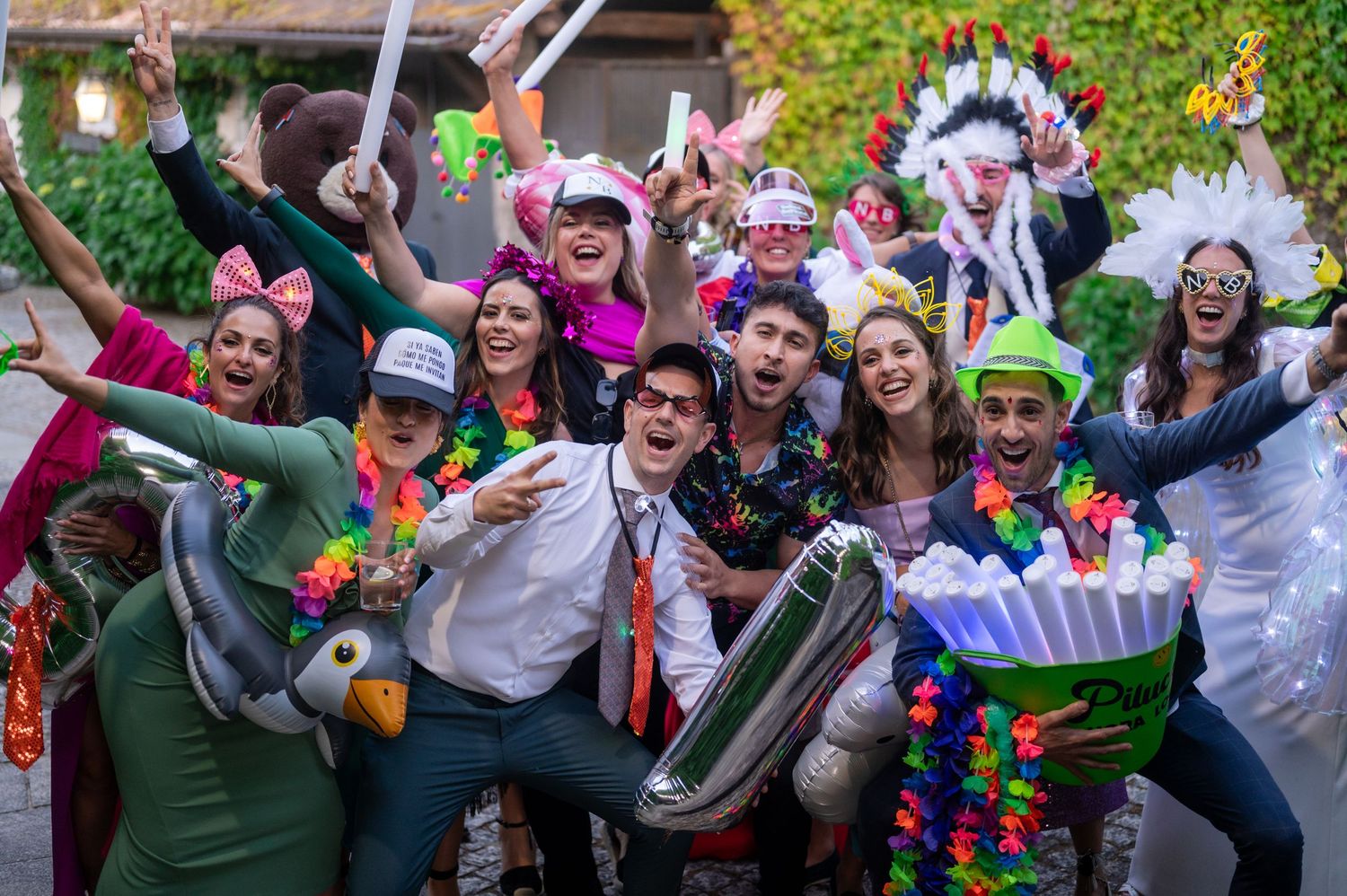 Large group of wedding guests jumping and celebrating with colorful props during an outdoor photo session.