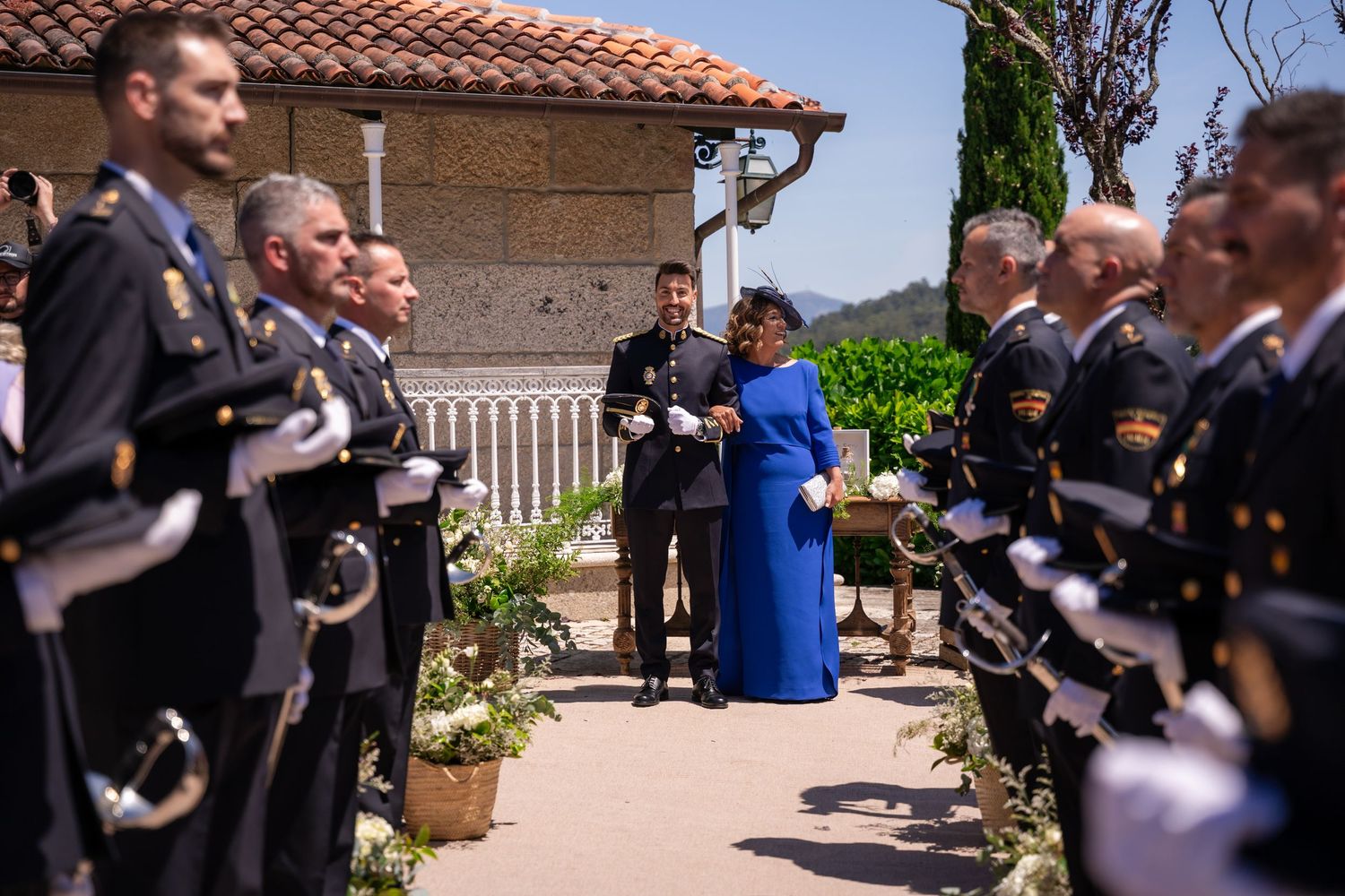 Military personnel in dress uniforms line up during an outdoor ceremony at a Mediterranean-style venue.