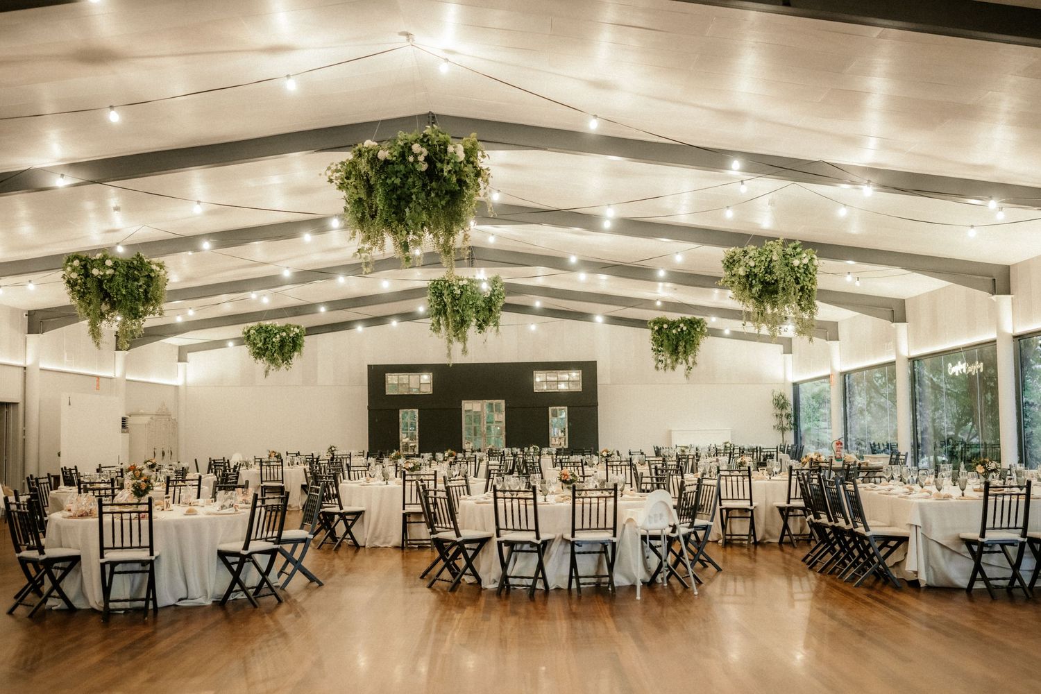 Elegant wedding reception venue with vaulted white ceiling, hanging greenery, and round tables with black chairs.