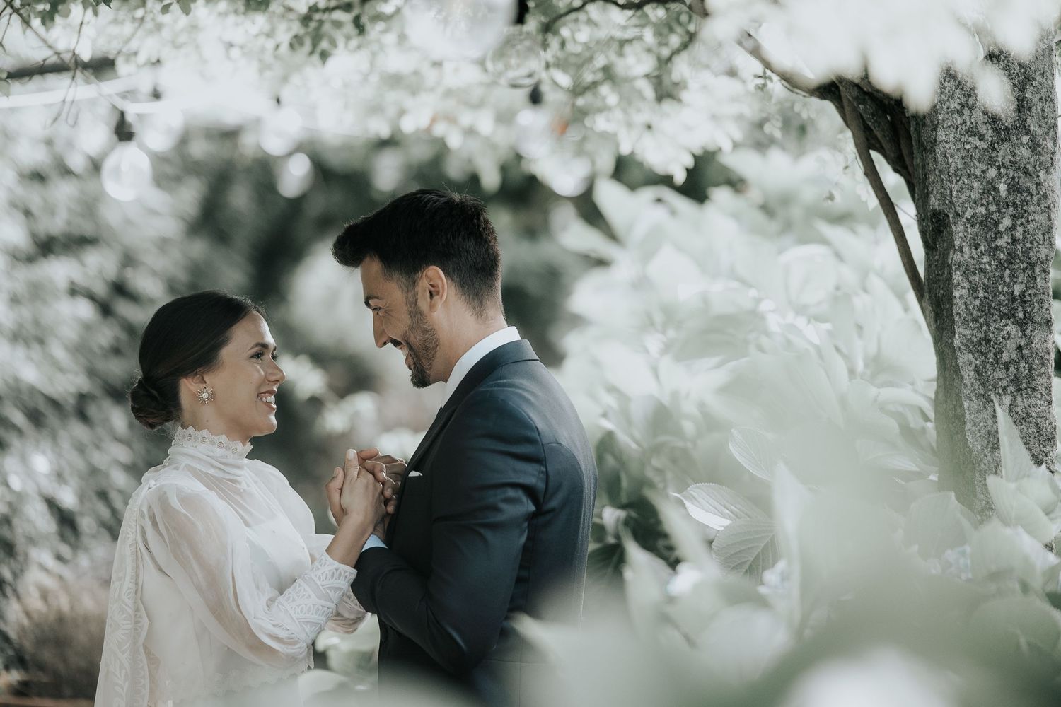 Couple in wedding attire share an intimate moment outdoors surrounded by soft white blossoms and foliage.