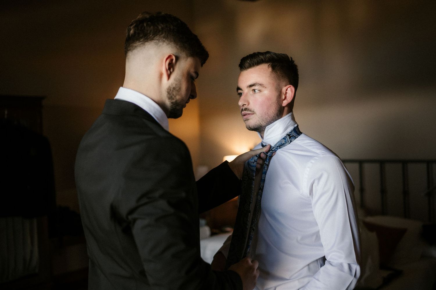 Candid wedding preparation scene shows men in formal wear adjusting ties and suits in warm indoor lighting.
