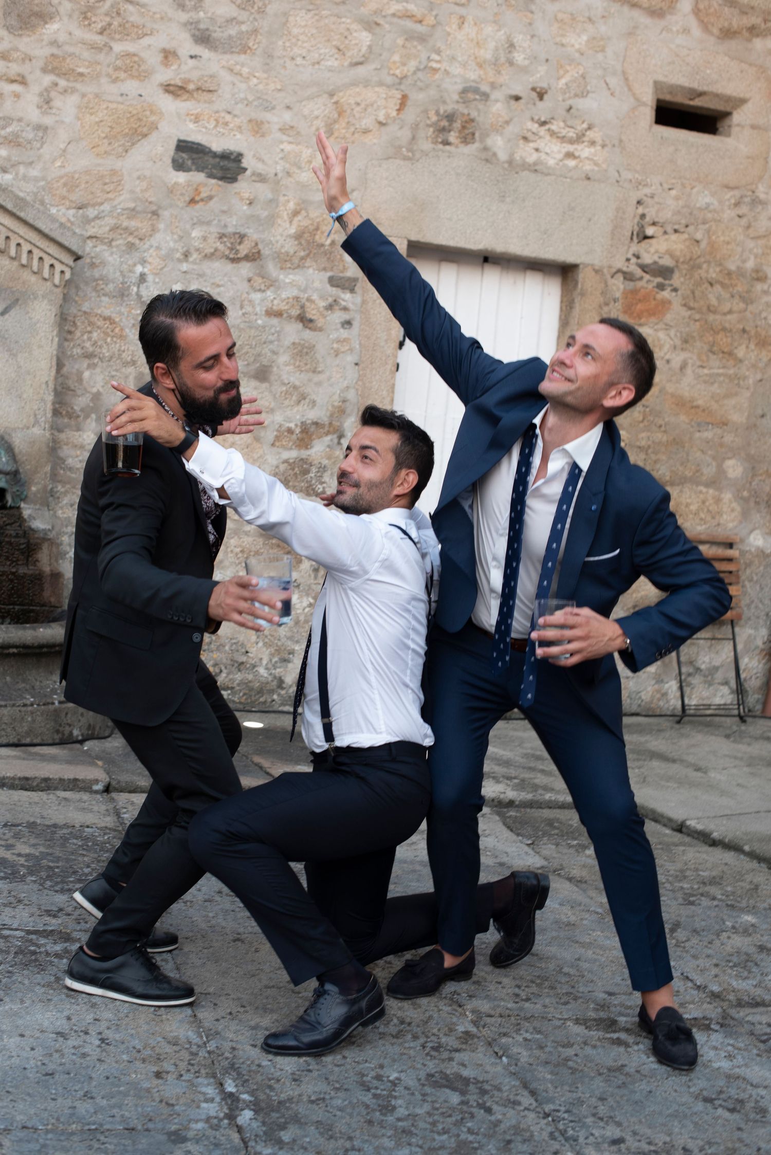Men in formal attire playfully reenact fighting moves against a stone wall backdrop.