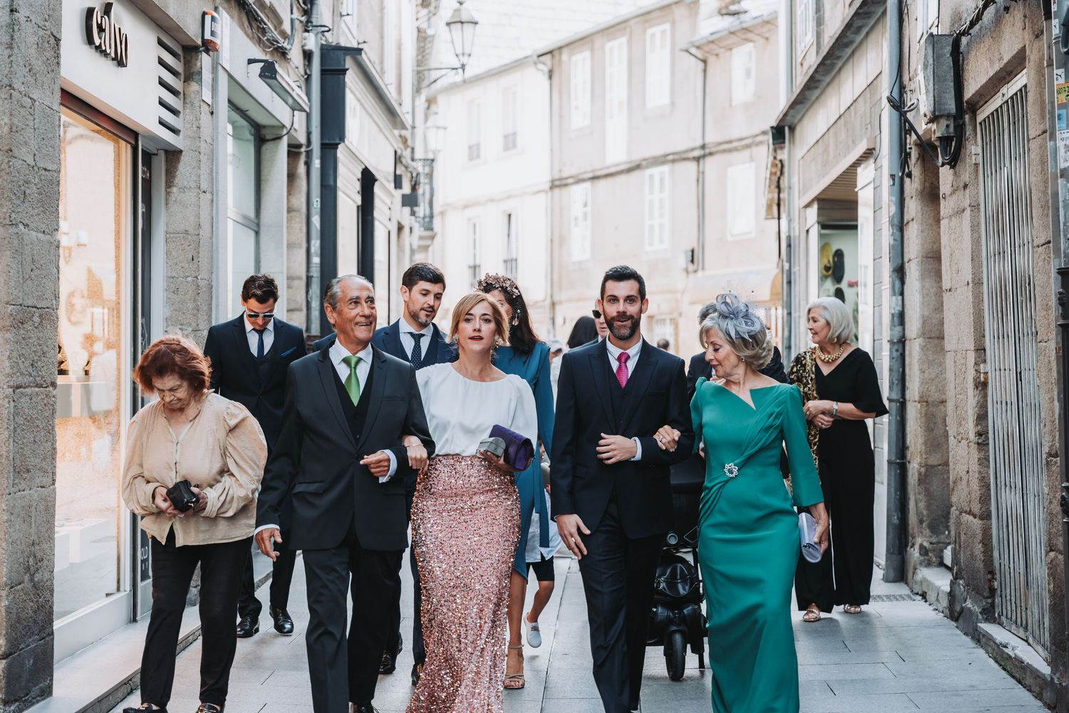 Elegant wedding party walks together through narrow European city street wearing formal attire in green and blush tones.