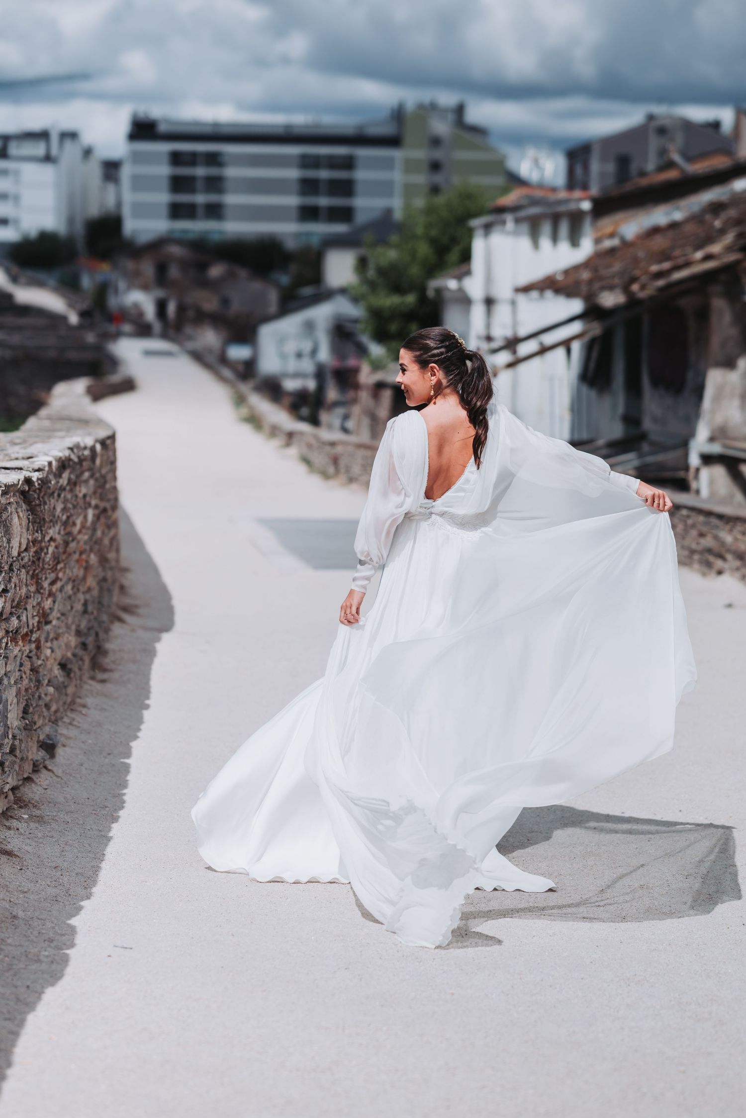 A white bridal gown creates dramatic movement as it flows in the breeze along a stone wall path.