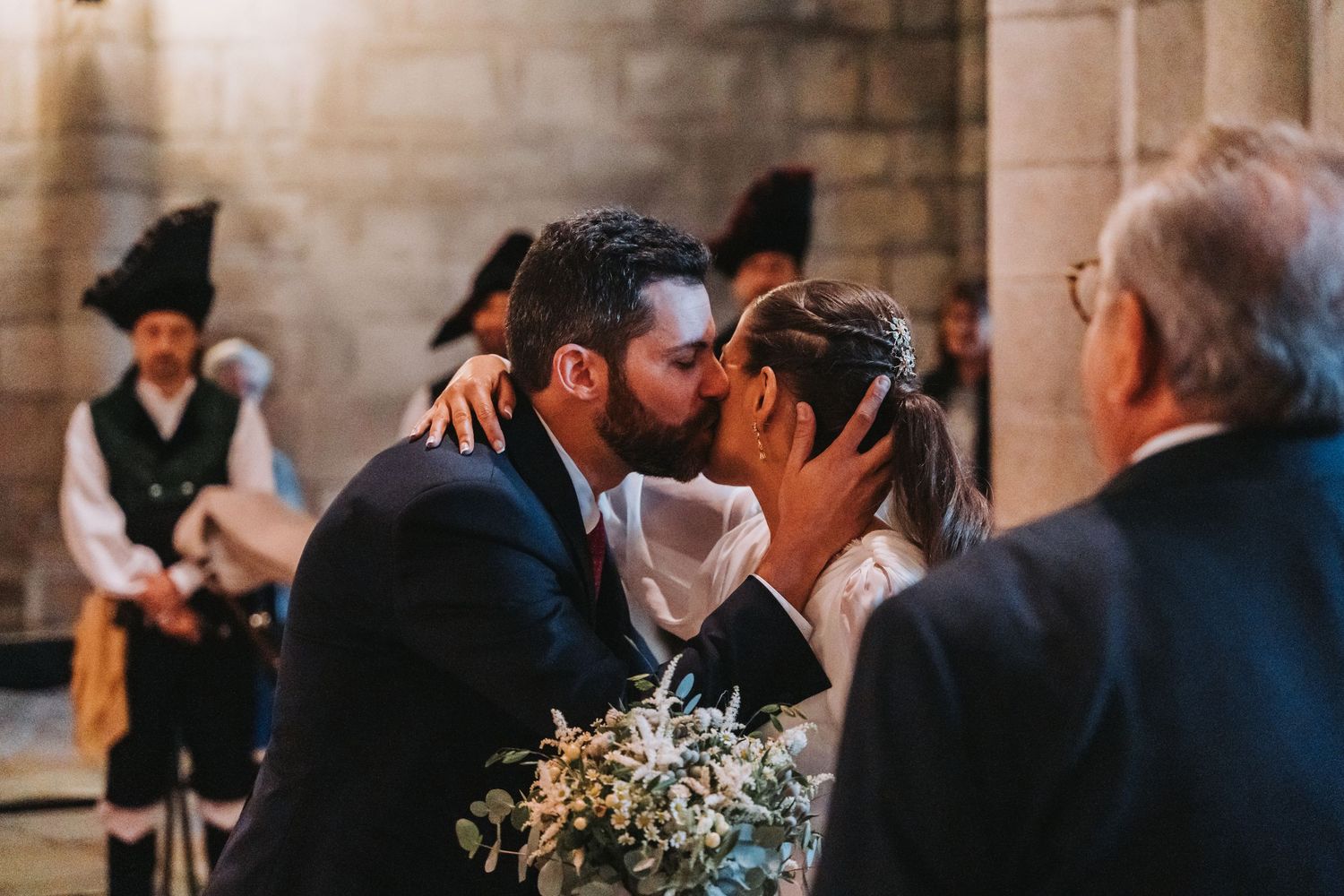 A couple shares a romantic kiss during their wedding ceremony in a dimly lit church setting.