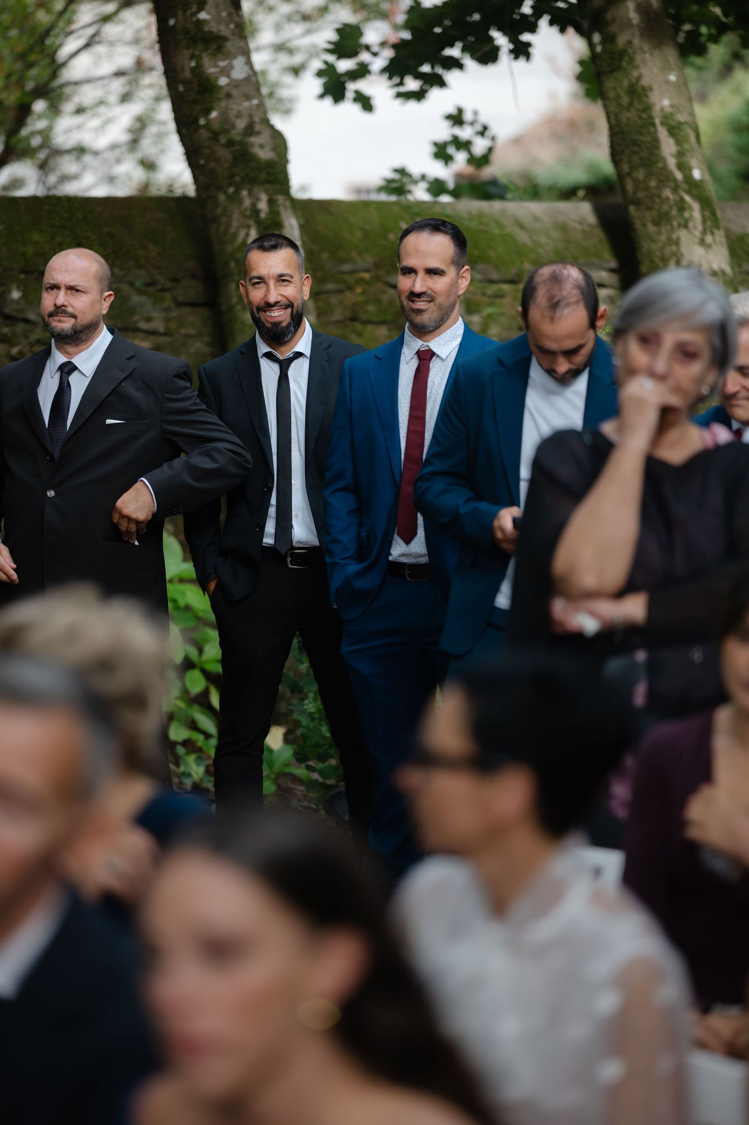 A group of men in formal suits and ties stand together outdoors near trees during what appears to be a social gathering.