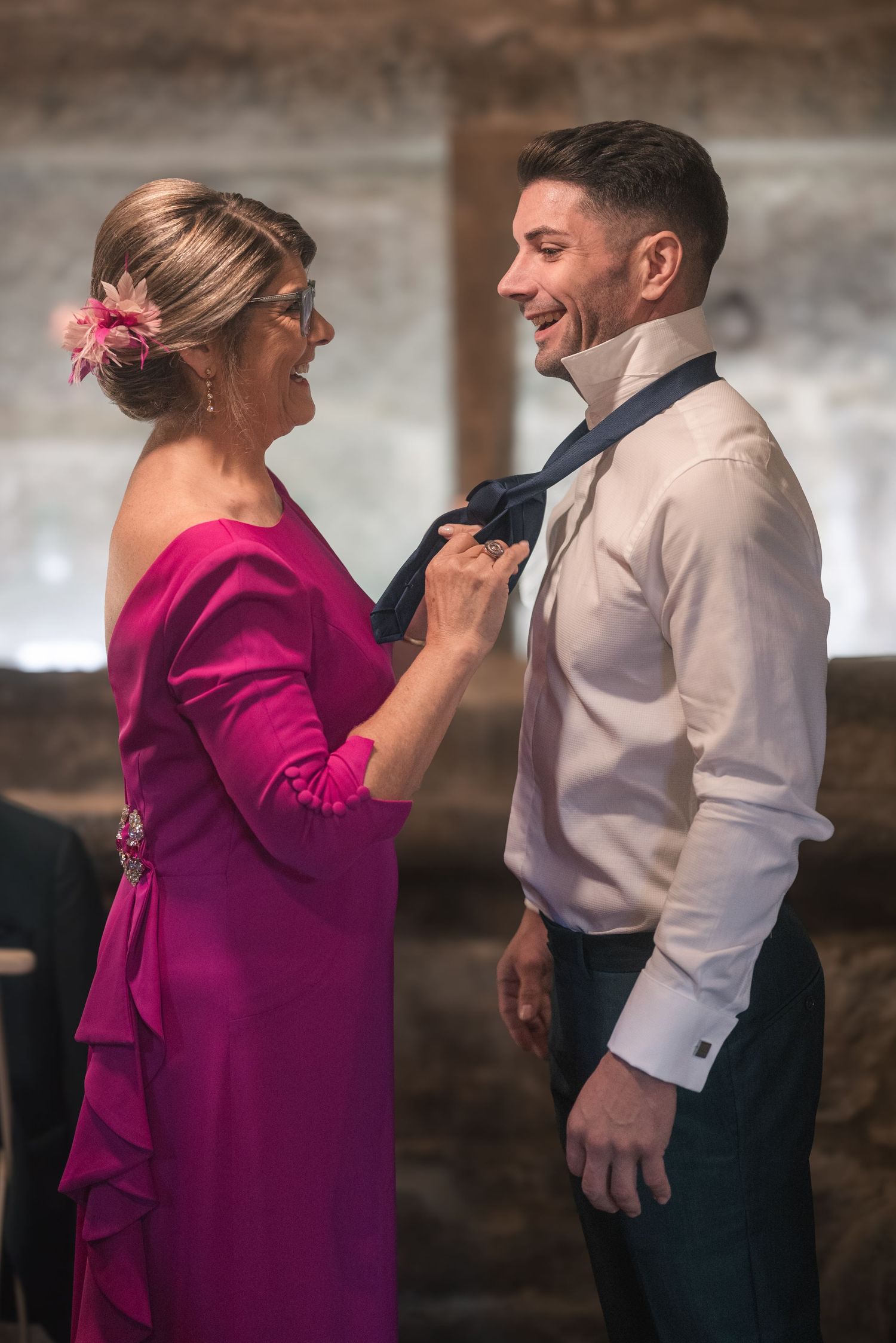 A woman in a magenta dress adjusts the tie of a man in a white dress shirt at a formal event.