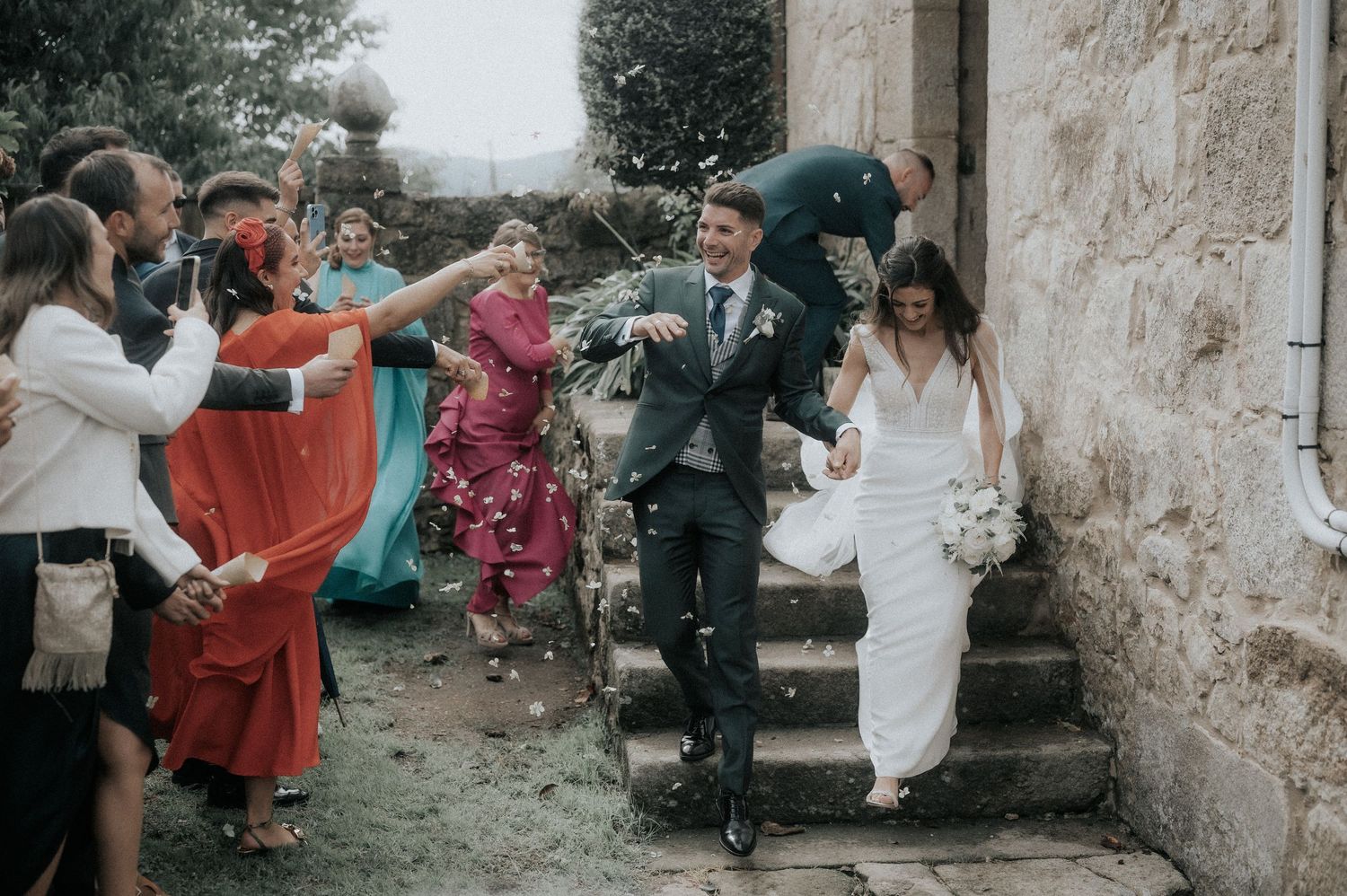 Wedding guests celebrate and throw confetti outside a rustic stone building.