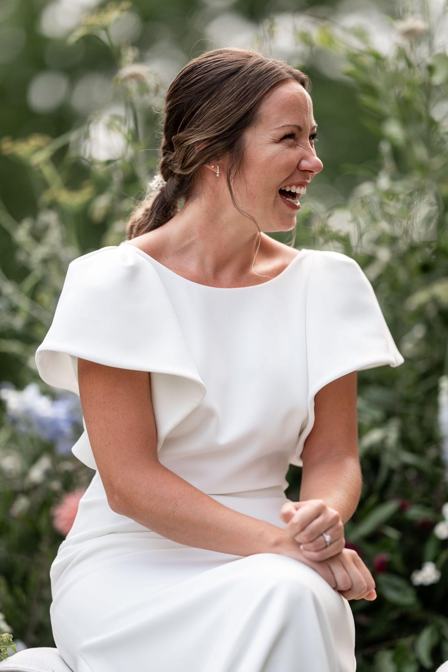 A joyful person in a white wedding dress laughs and spins in an outdoor garden setting.