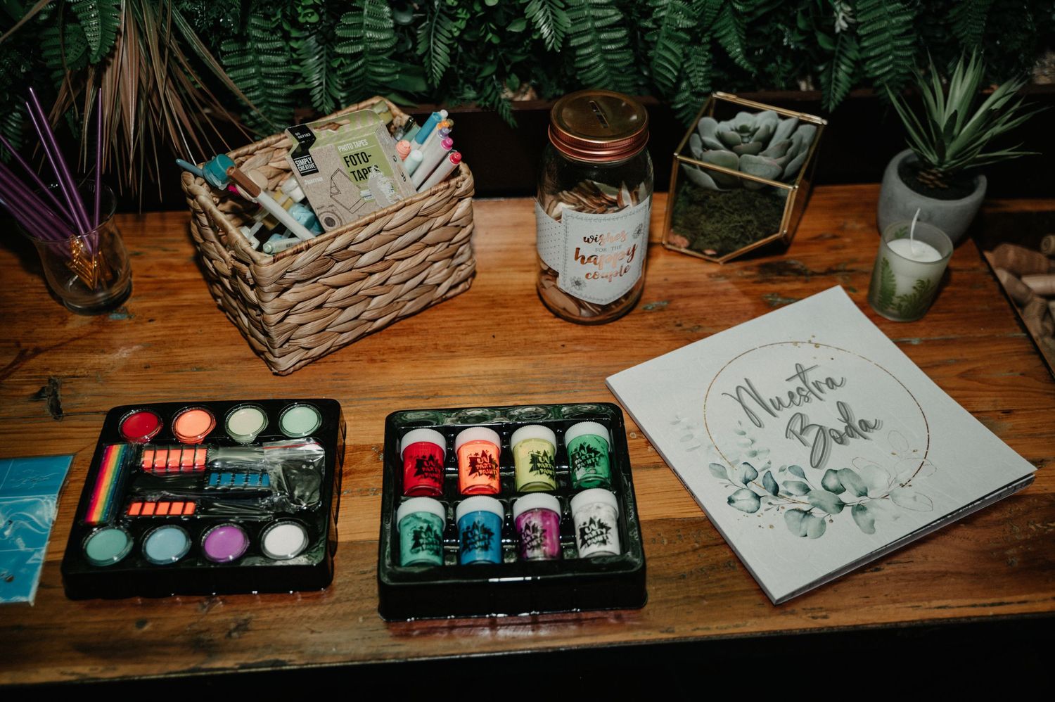 Colorful ink bottles and art supplies arranged on a wooden table with plants and a mason jar in the background.