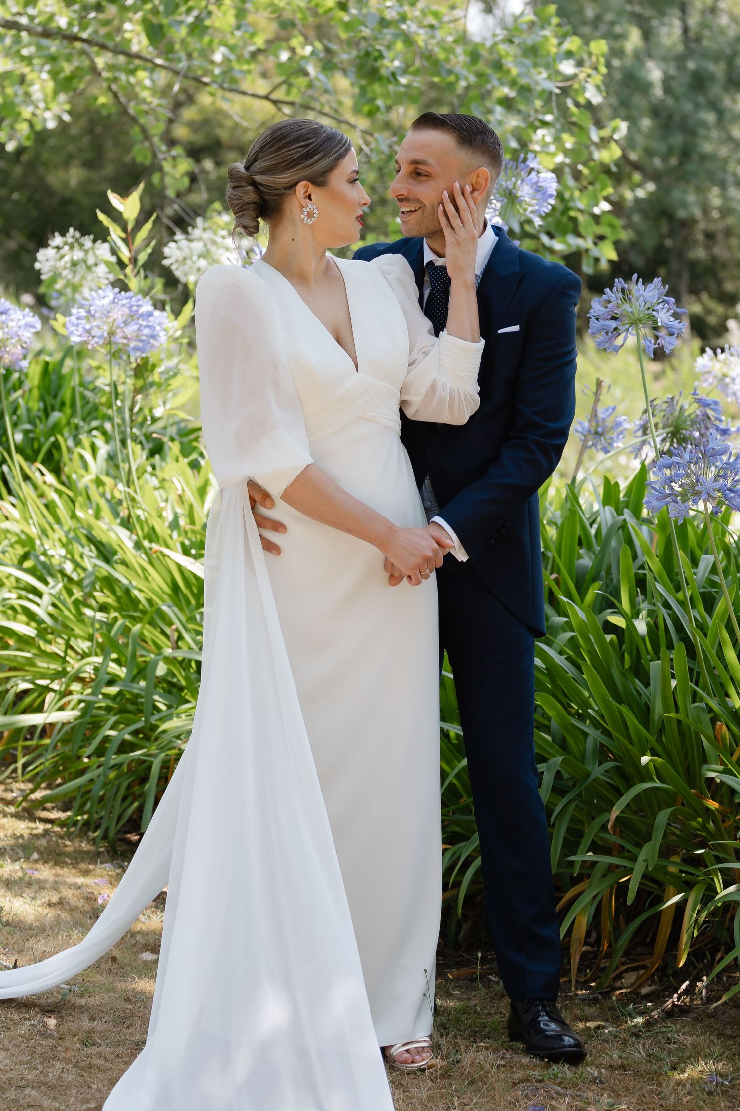 Romantic wedding couple poses together in a garden setting with blooming purple agapanthus flowers in the background.