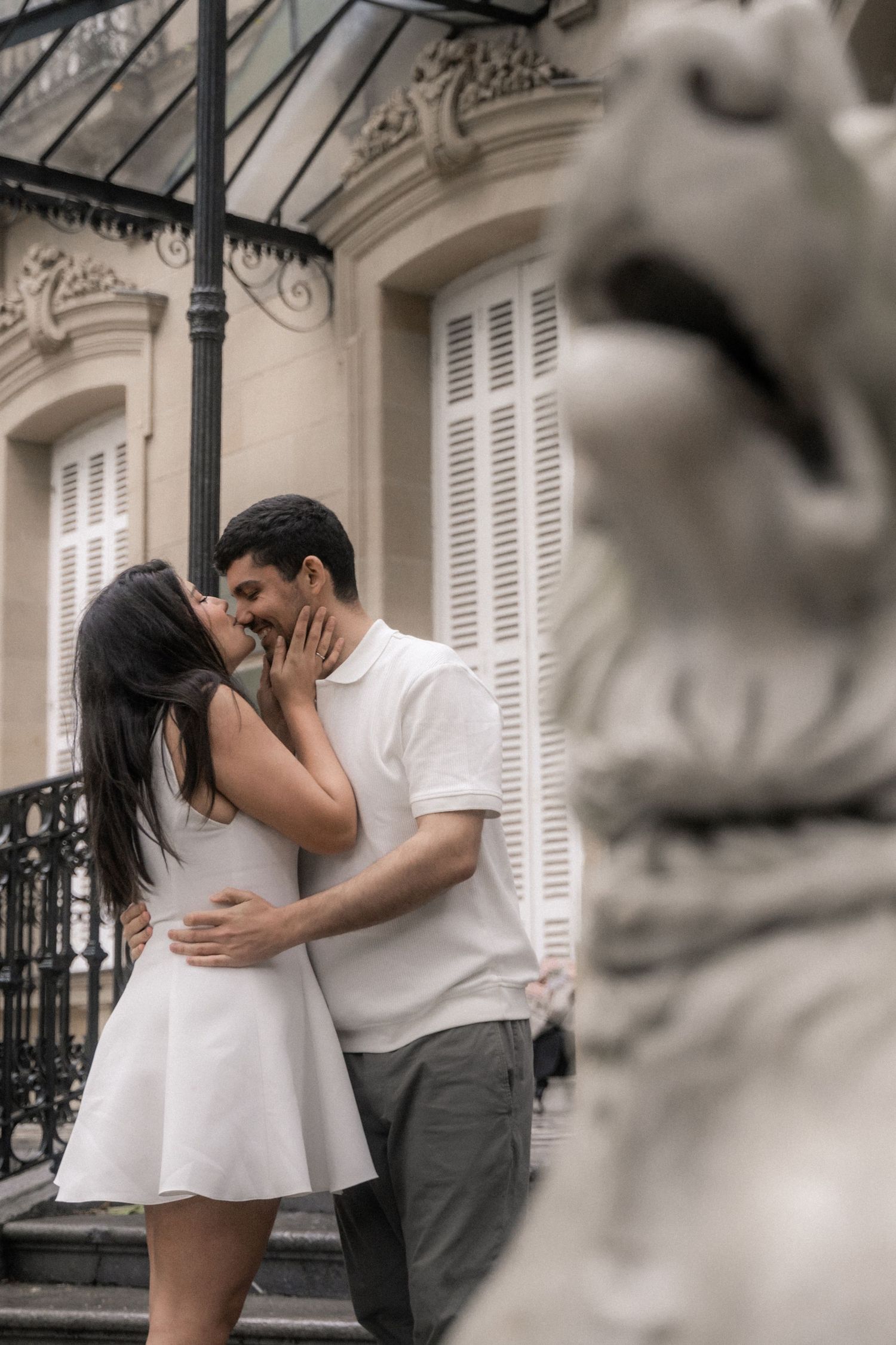 Romantic couple embraces and kisses against a classic European building facade with white shuttered windows.