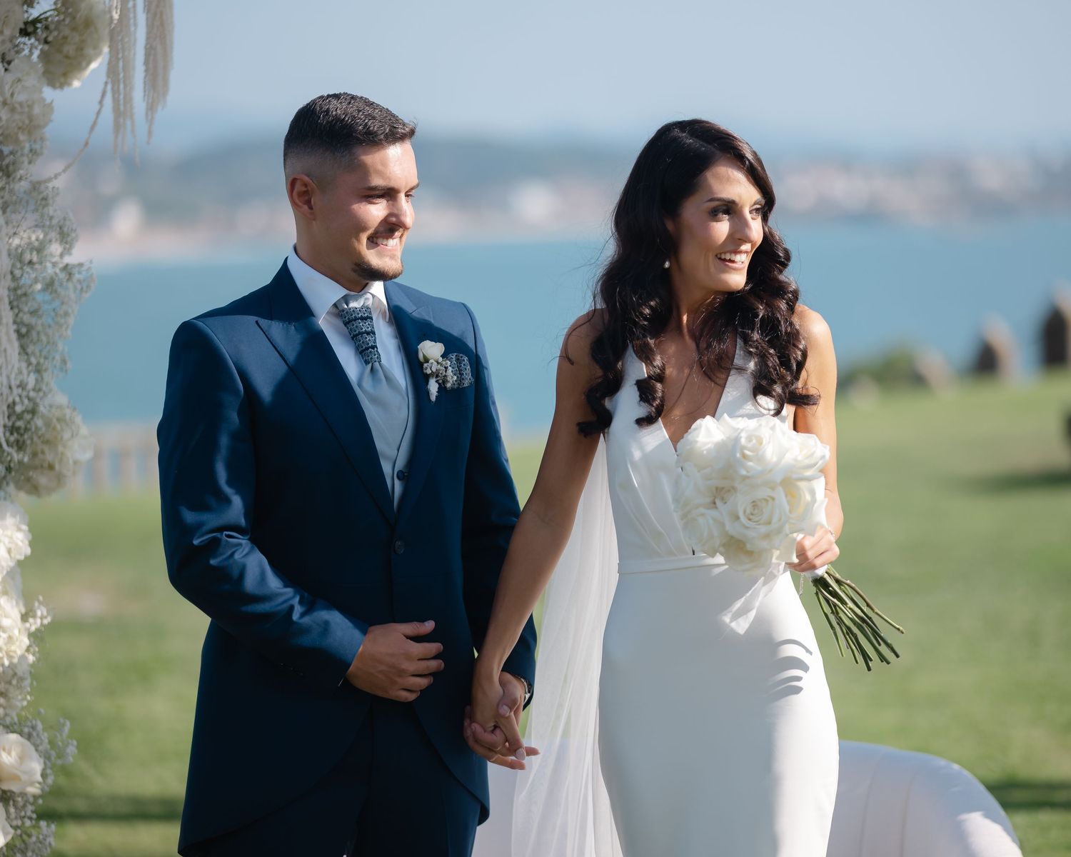 A bride and groom hold hands during their outdoor wedding ceremony overlooking a scenic waterfront view.