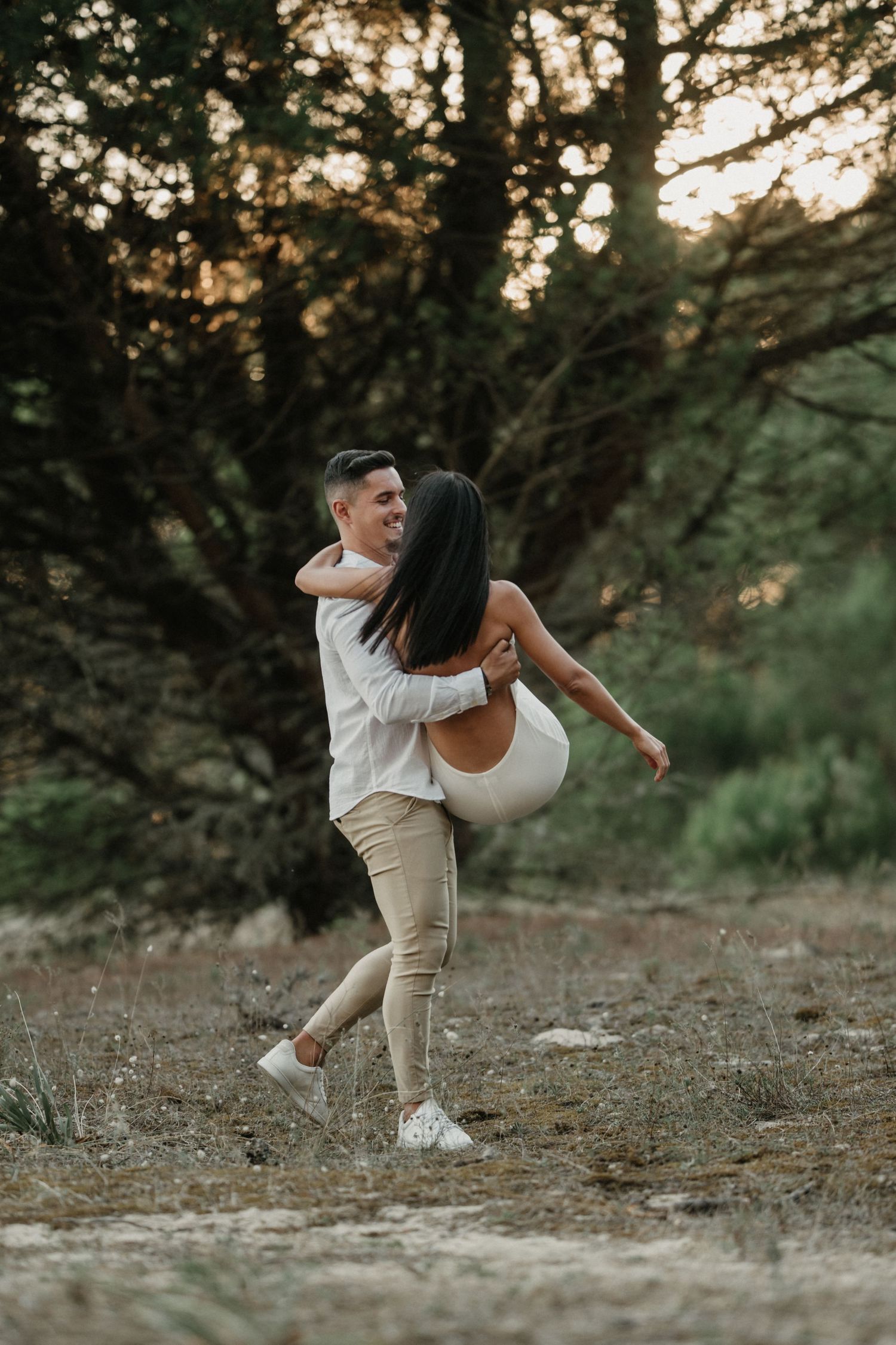 A couple in white and beige outfits dances playfully in a natural outdoor setting at sunset.