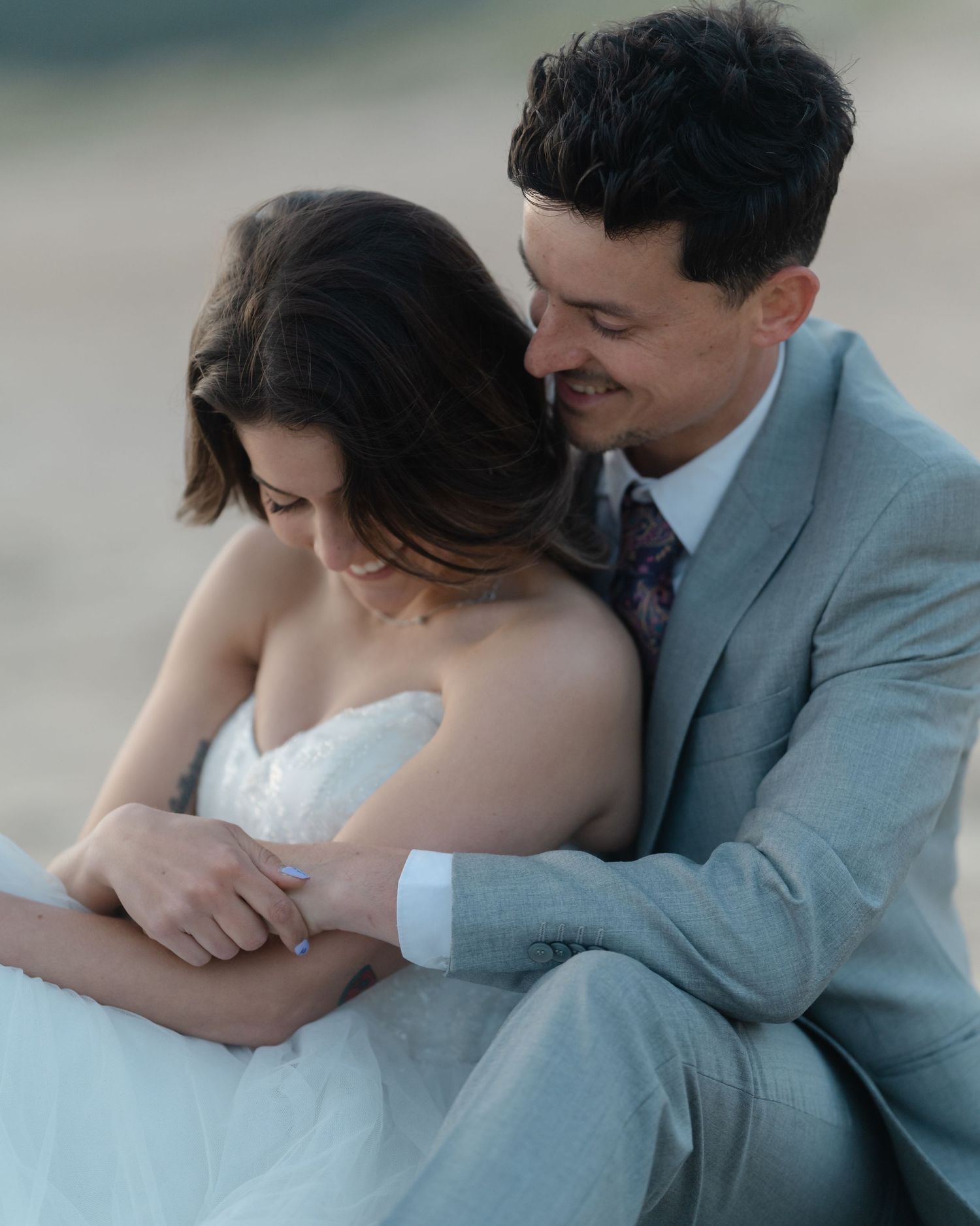 A smiling groom in a gray suit embraces his bride in a white strapless wedding dress as they sit together outdoors.