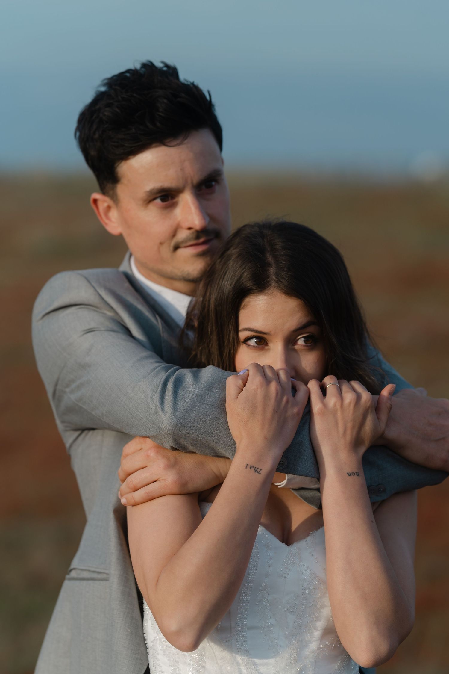 A groom in a gray suit embraces his bride from behind as she playfully hides her smile during outdoor wedding photos.