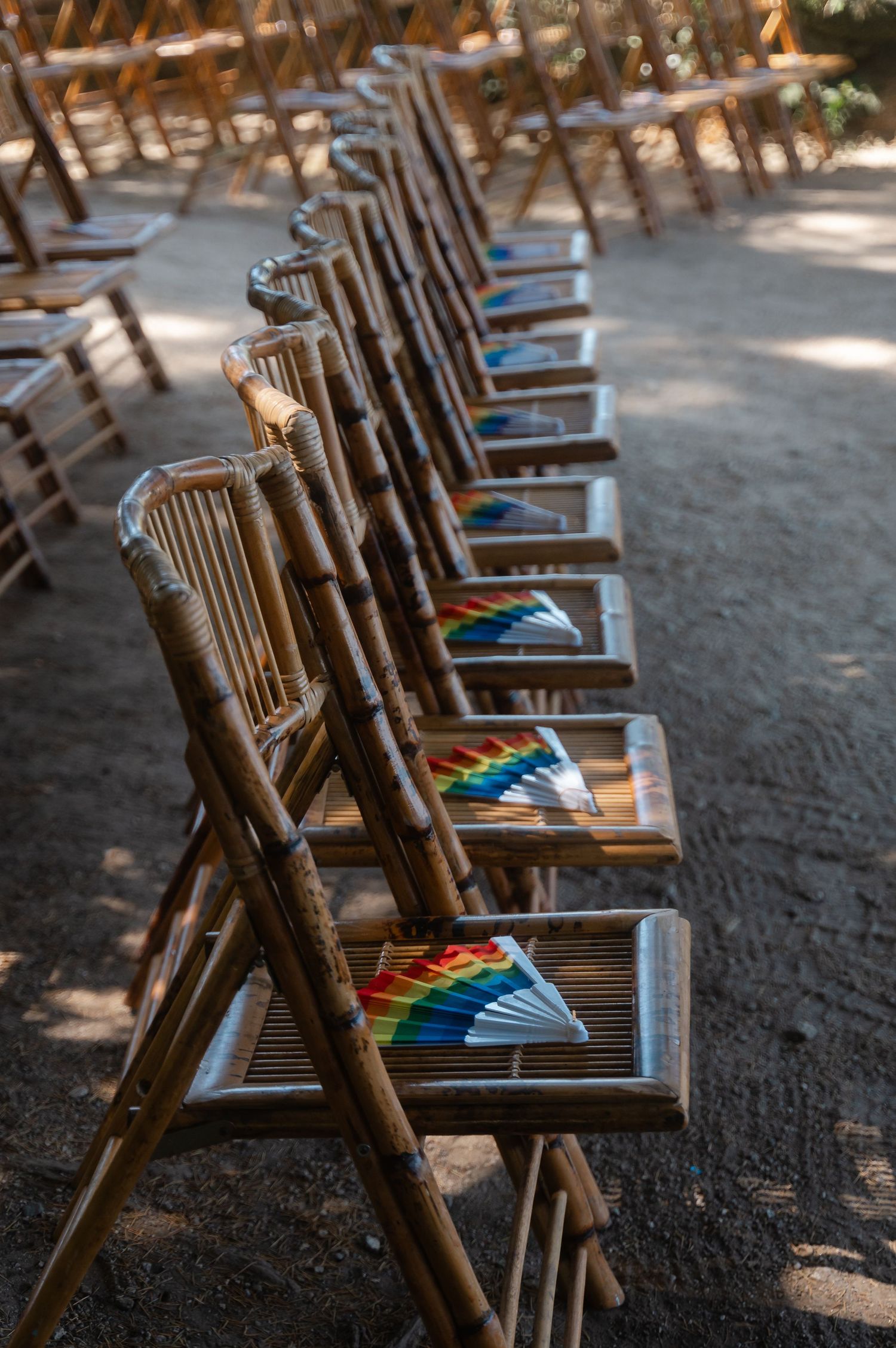 Wooden rocking chairs lined up in a row with colorful fans placed on their seats in dappled sunlight.