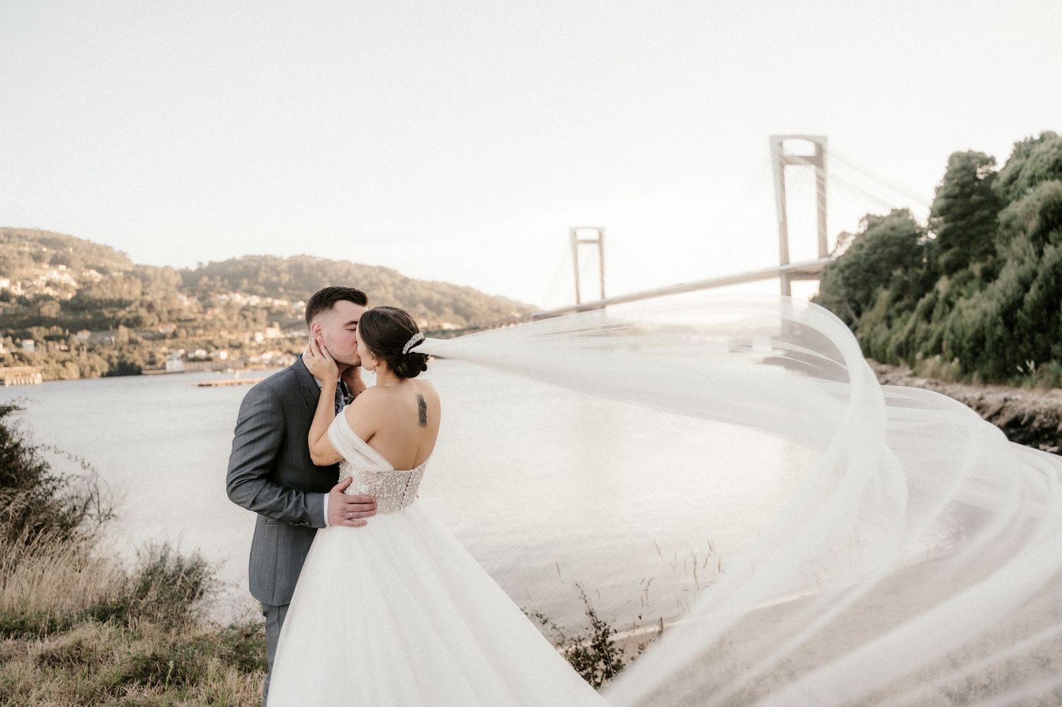 A wedding couple embracing with a flowing veil against a scenic bridge backdrop at sunset.