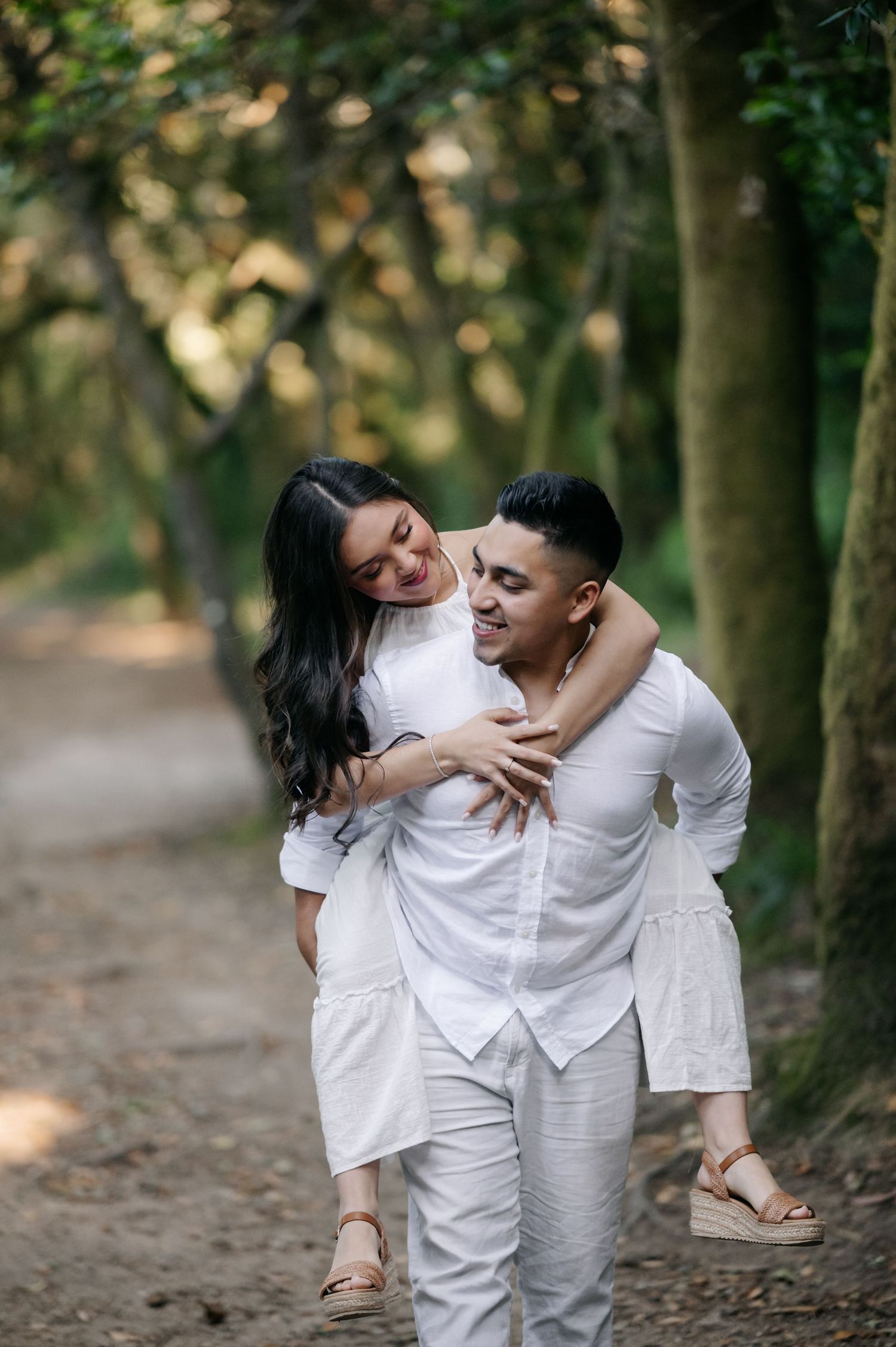 A playful couple dressed in white laughs together while embracing on a forest trail.