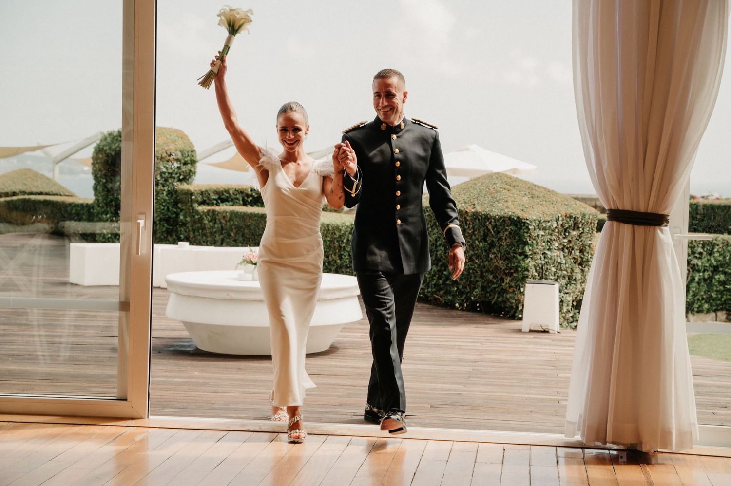 A couple dances together on a wooden floor in an elegant venue with flowing white curtains.