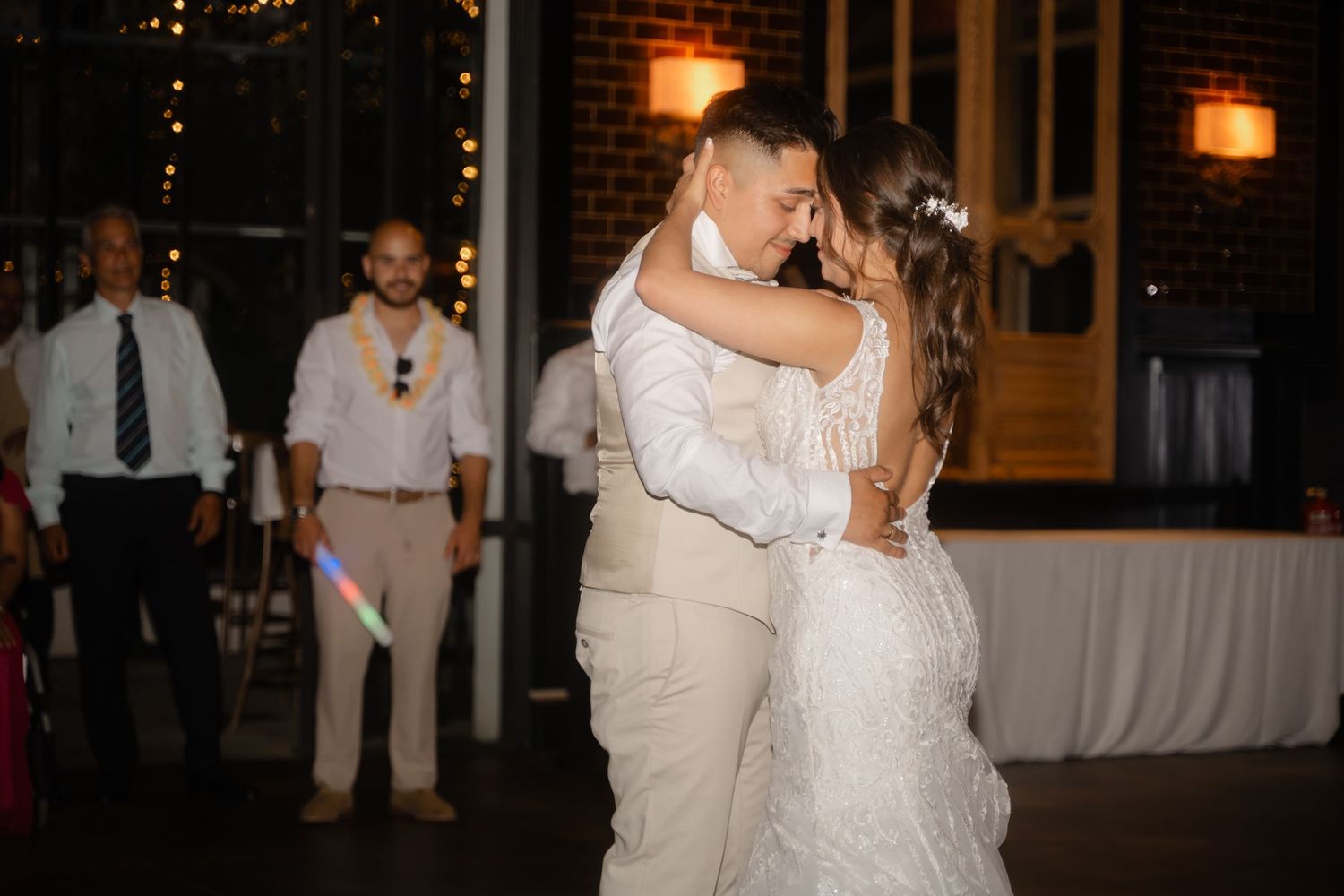 A couple shares a romantic dance moment at a wedding reception with warm lighting and brick walls in the background.