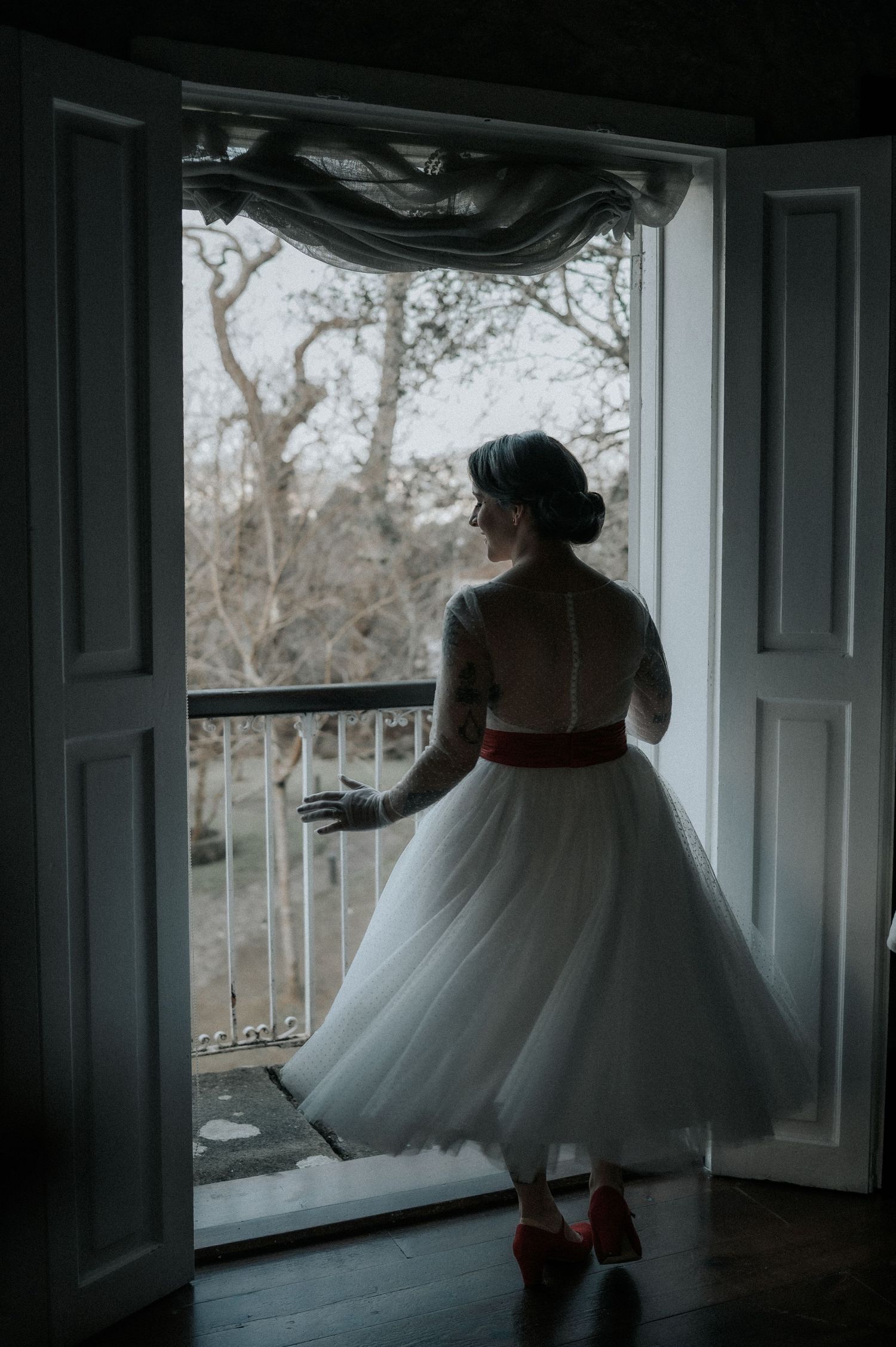 A silhouetted figure in a white tulle dress stands by a window in moody, dramatic lighting.