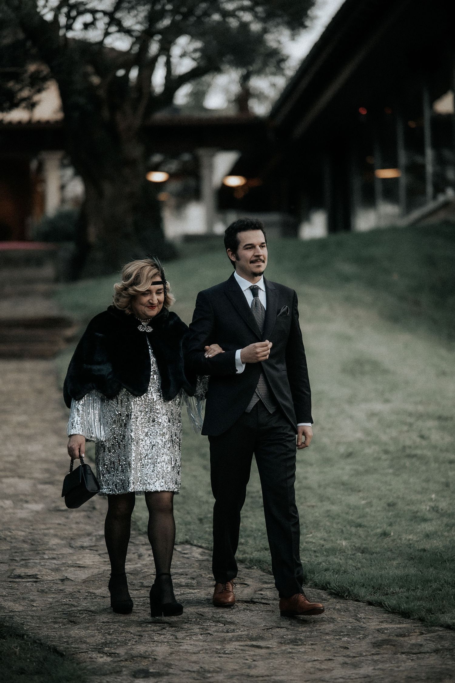A couple in formal attire walking together on a path during a moody outdoor photoshoot.