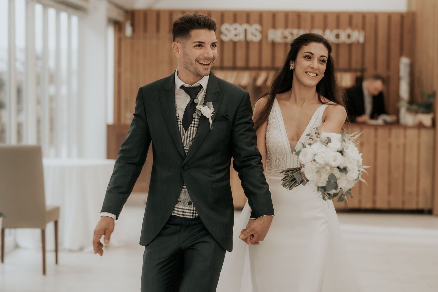 A bride and groom walk together in a rustic setting with wooden panels in the background.
