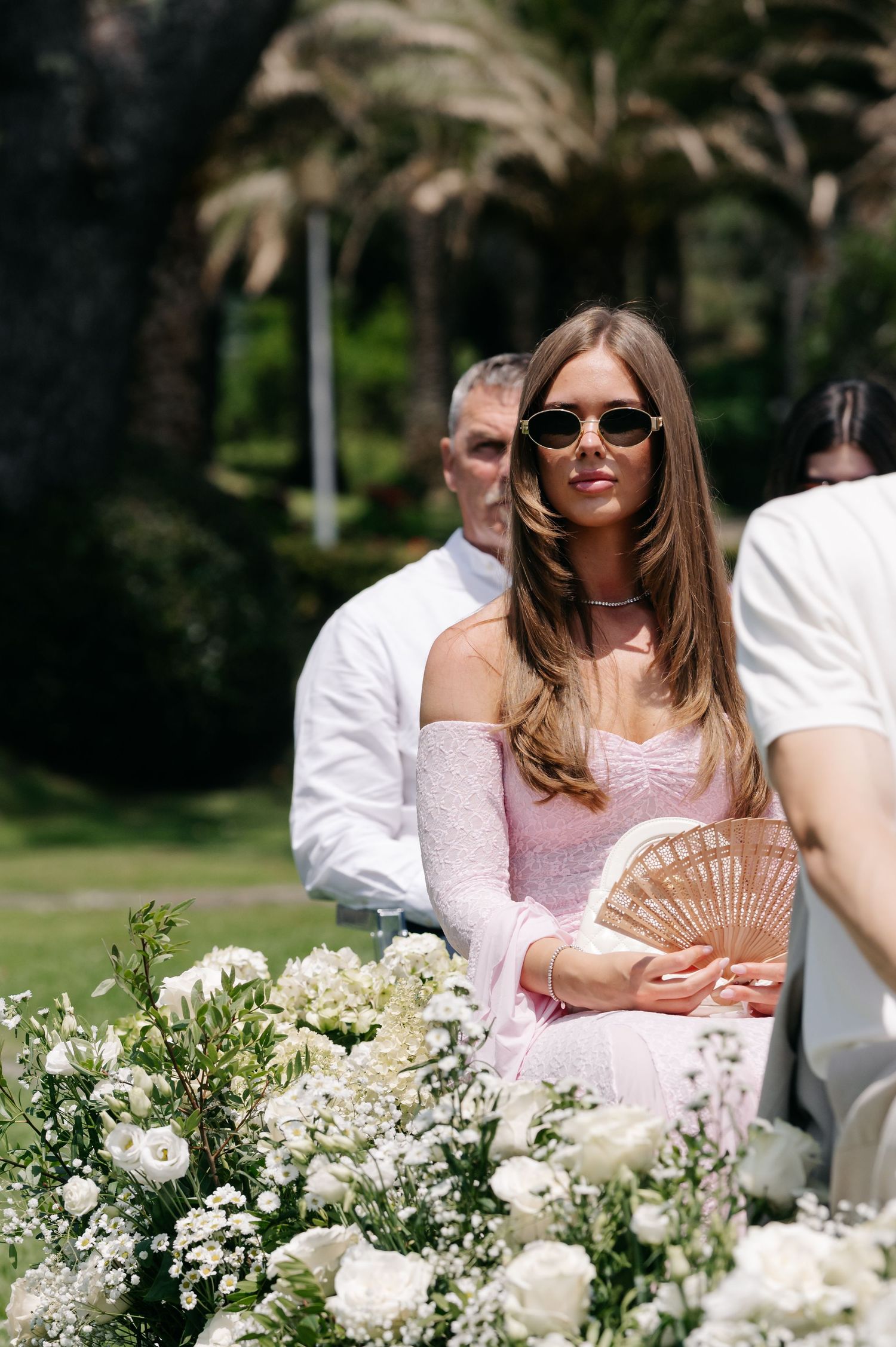 Wedding guest in pink dress sits at outdoor reception table decorated with elegant white floral arrangements.