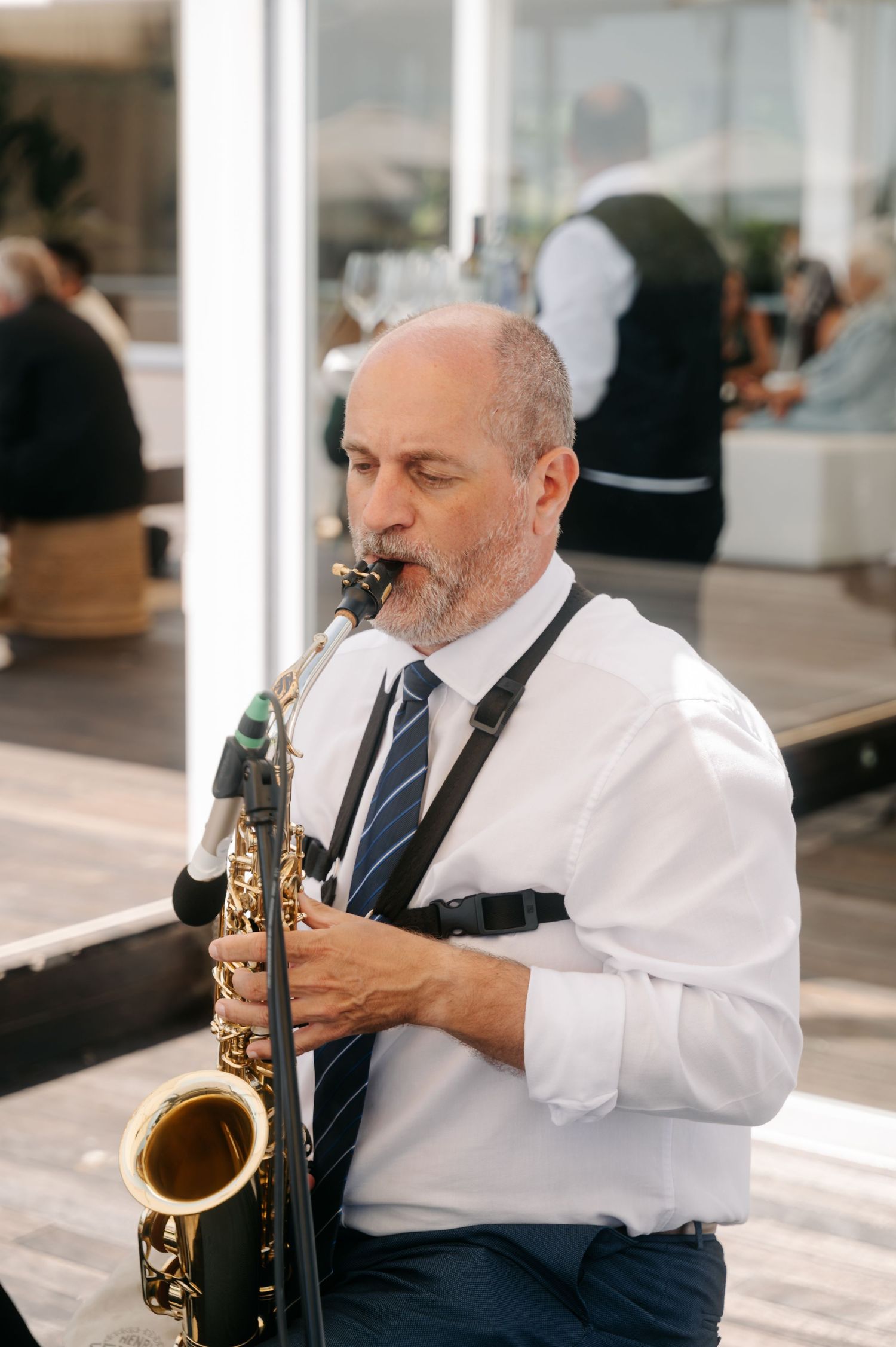 A saxophonist in a white shirt and dark tie performs in a sunlit room with large windows.
