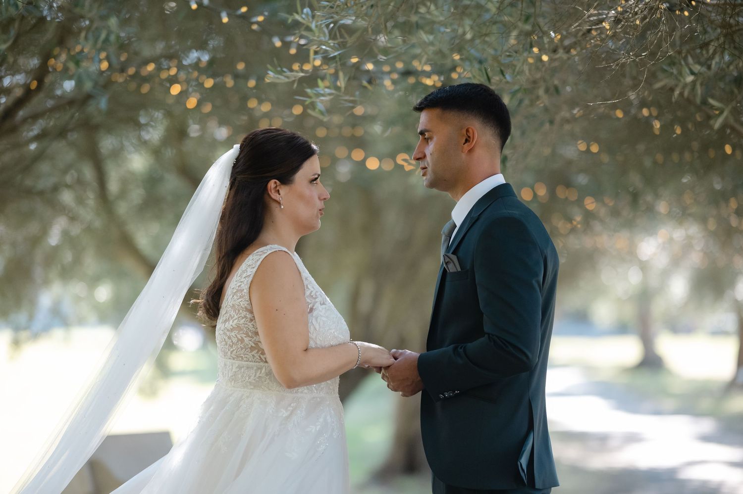 A bride and groom share an intimate moment holding hands in a dreamy outdoor setting with soft sunlit trees.