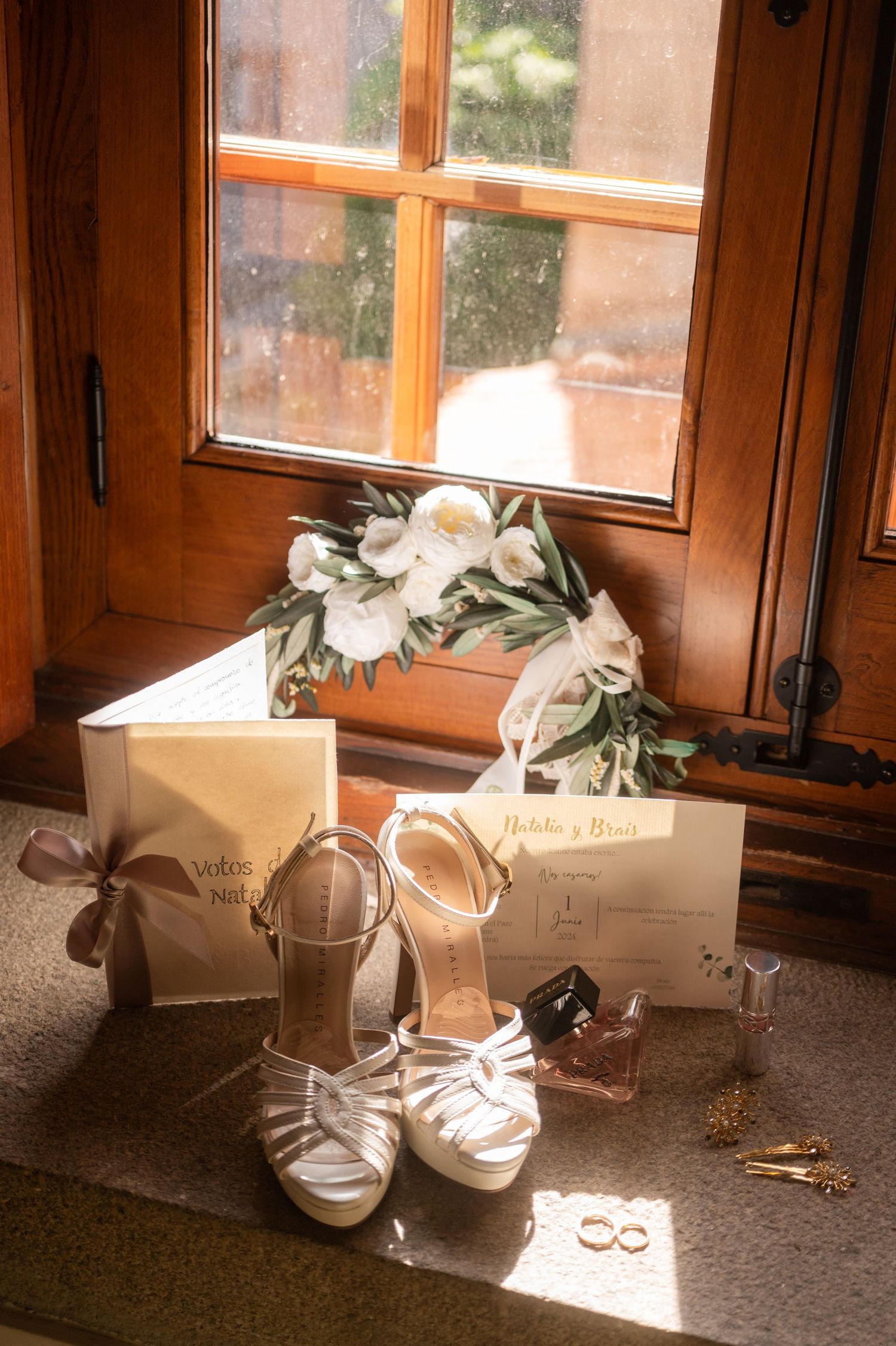 Wedding accessories including silver heels, floral crown, and invitation cards arranged by a wooden window in warm light.