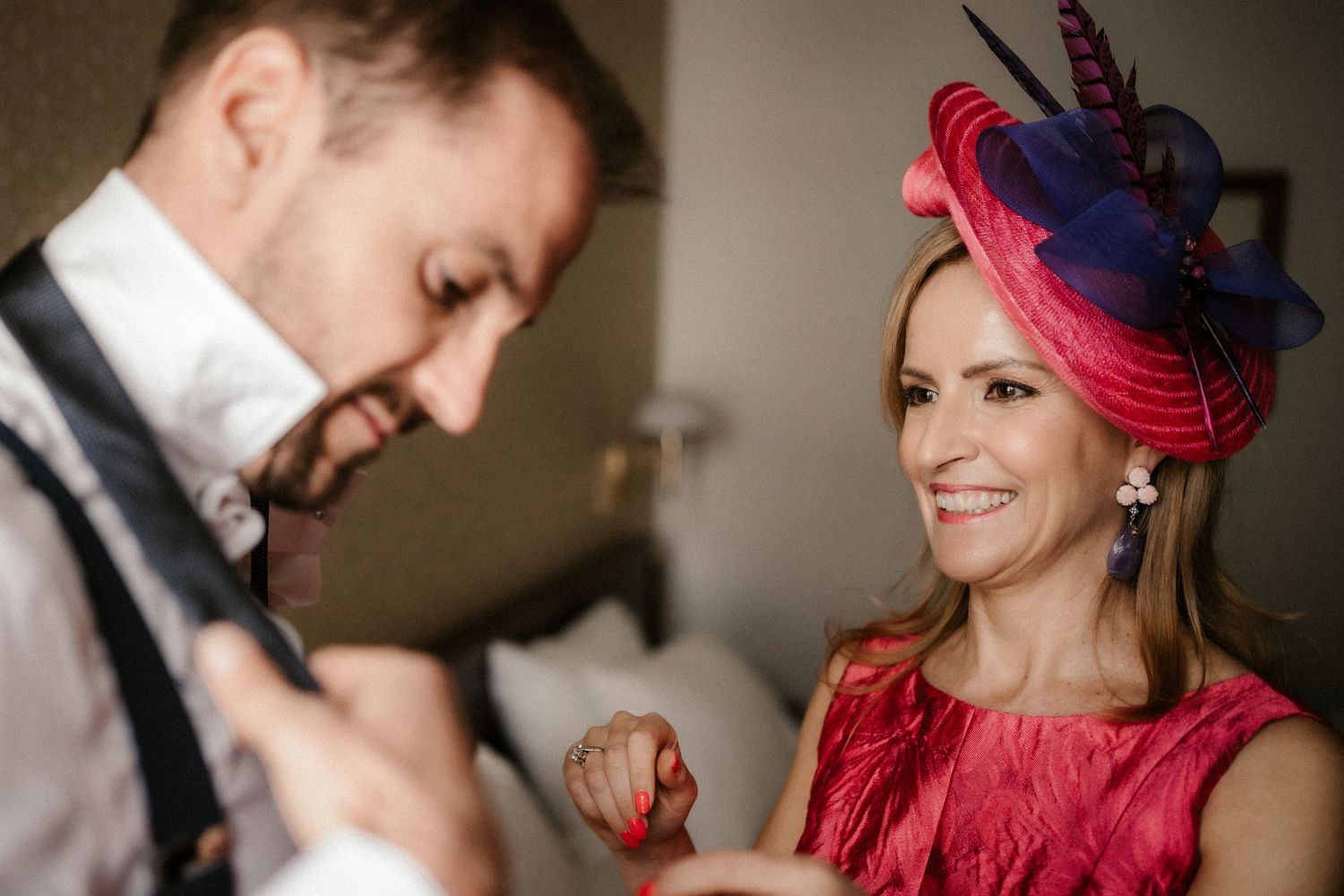 A couple shares a joyful moment at a formal event, with a vibrant red dress and fascinator adding elegant flair.