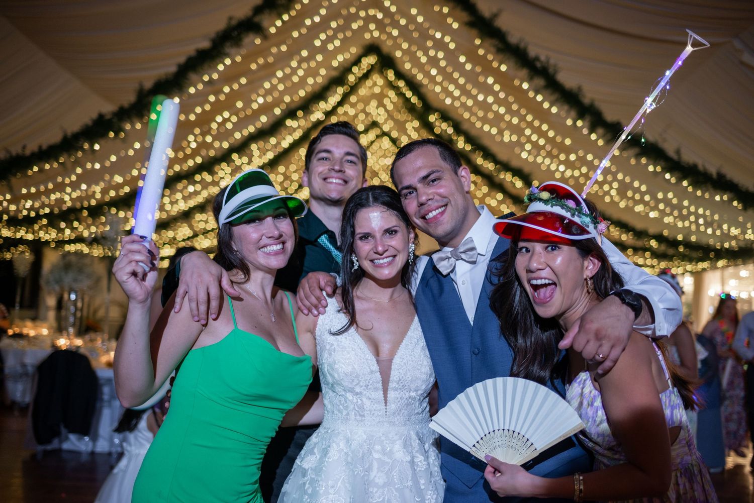Wedding party celebrates with light-up wands under a canopy of golden string lights.