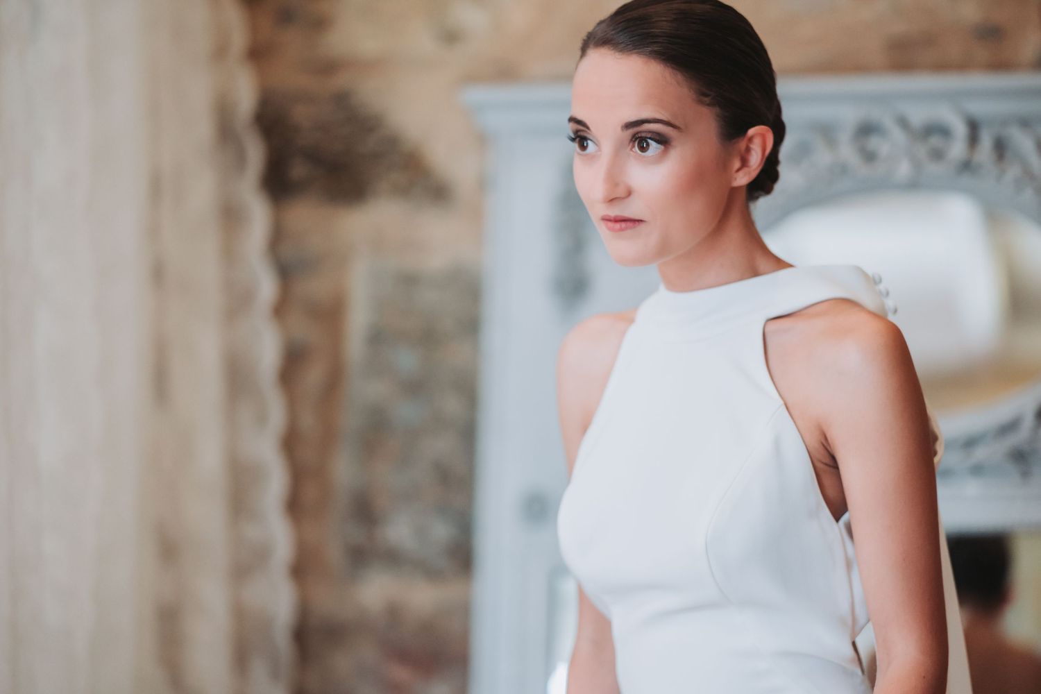 Elegant figure in a sleek white sleeveless dress poses in front of a rustic stone wall with soft lighting.