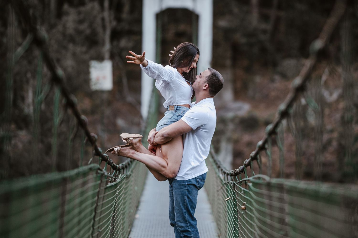Two people embrace while standing on a wooden footbridge in a forest setting.