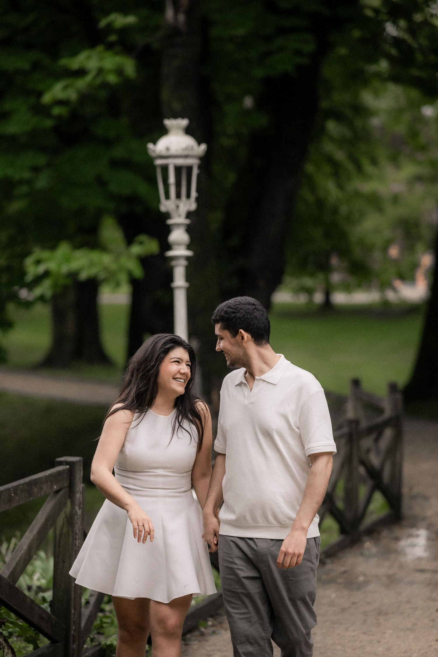 A couple in white and grey outfits share romantic moments while leaning on a wooden railing in a lush park setting.