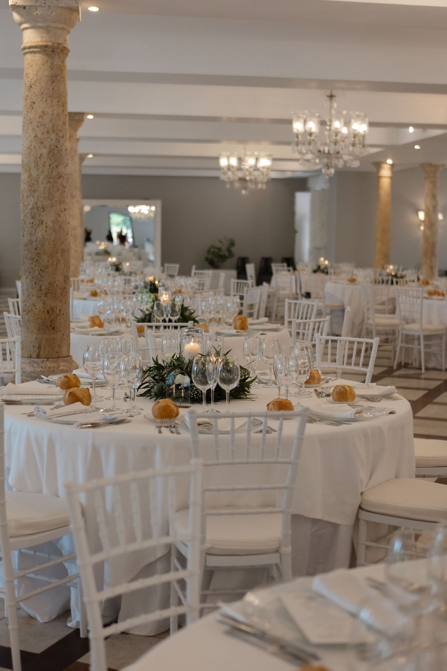 Elegant wedding reception venue with crystal chandeliers, white tablecloths, and ornate stone columns in a bright banquet hall.