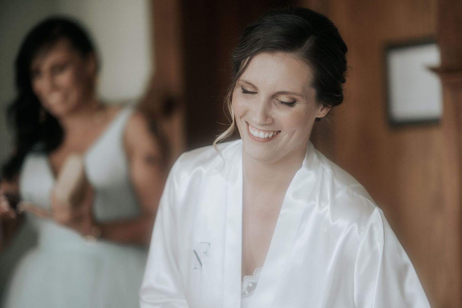 A bride in a white wedding dress smiles while getting ready in a warmly lit room.
