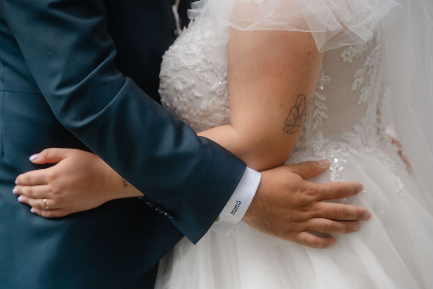 A close-up detail shot of hands embracing during a wedding, with a white lace dress and navy suit visible.