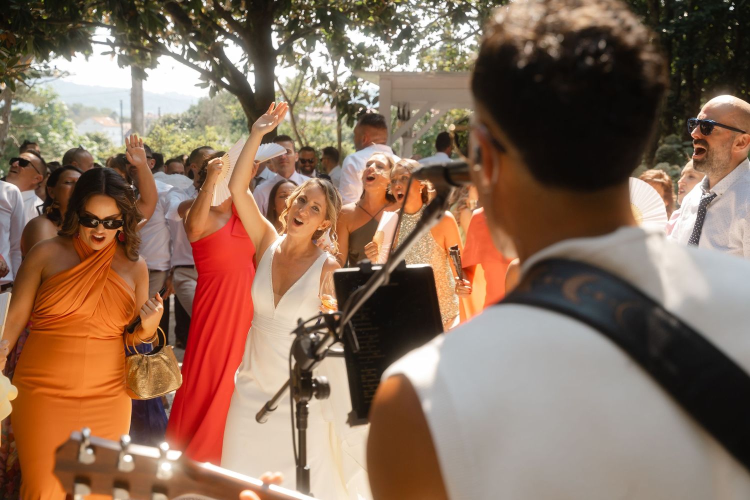 Wedding guests gathered outdoors under trees during a sunny daytime celebration.