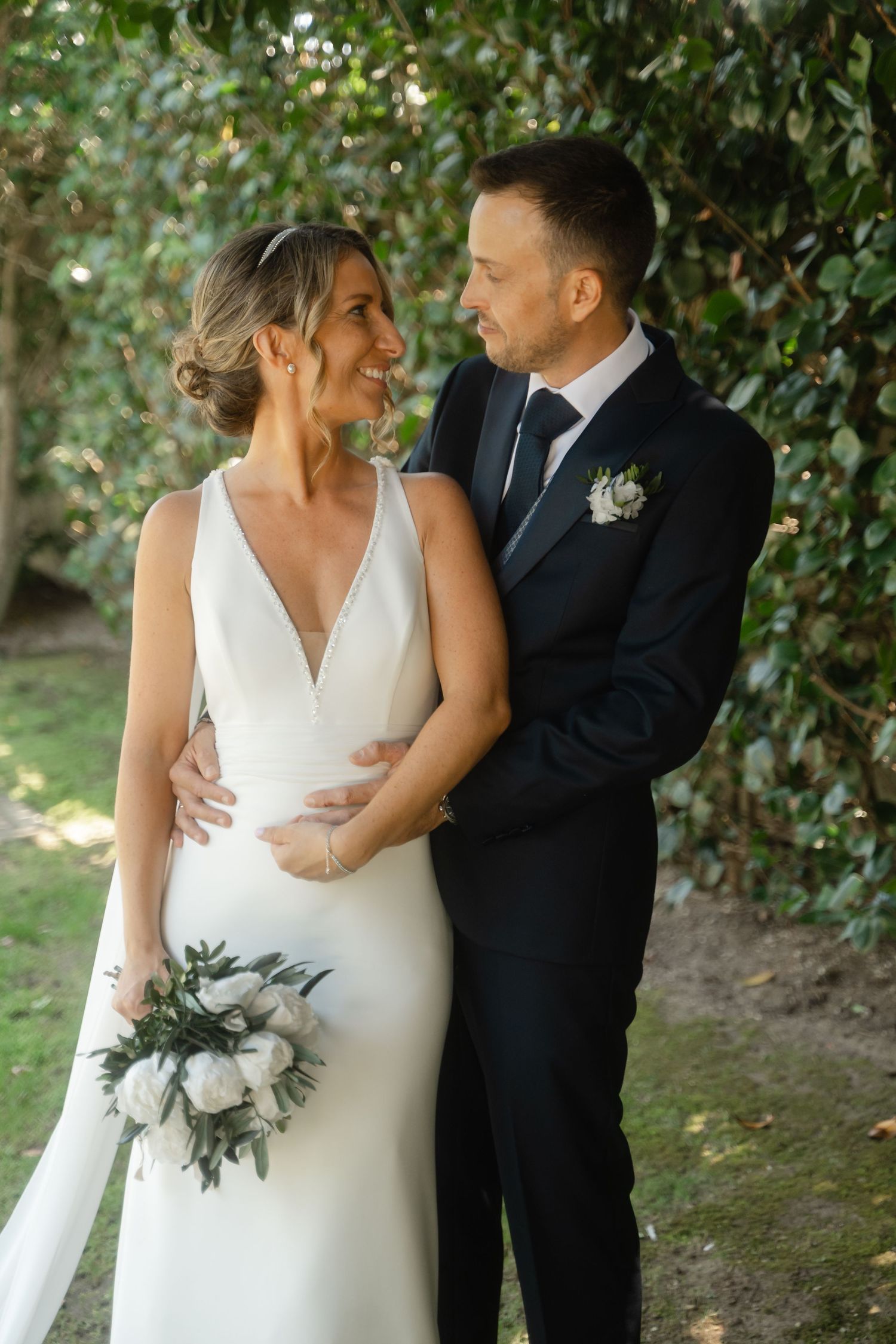 A bride in a sleek white dress and groom in dark suit share an intimate moment among garden foliage.