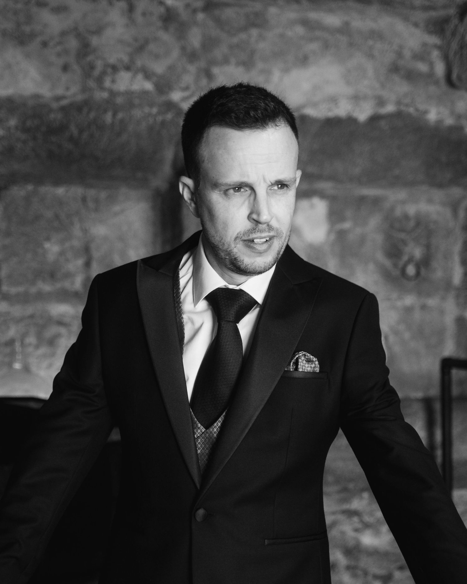 Black and white portrait of someone wearing a formal black tuxedo suit with pocket square against stone wall.