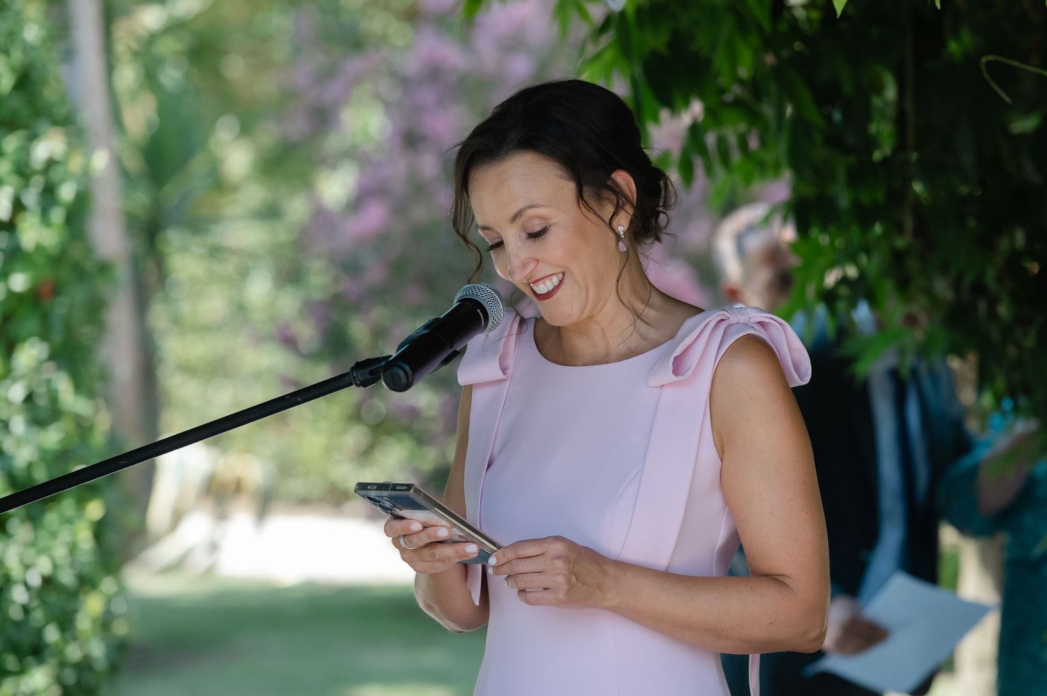 A person in a light dress sits outdoors in a garden setting while looking at their mobile phone.